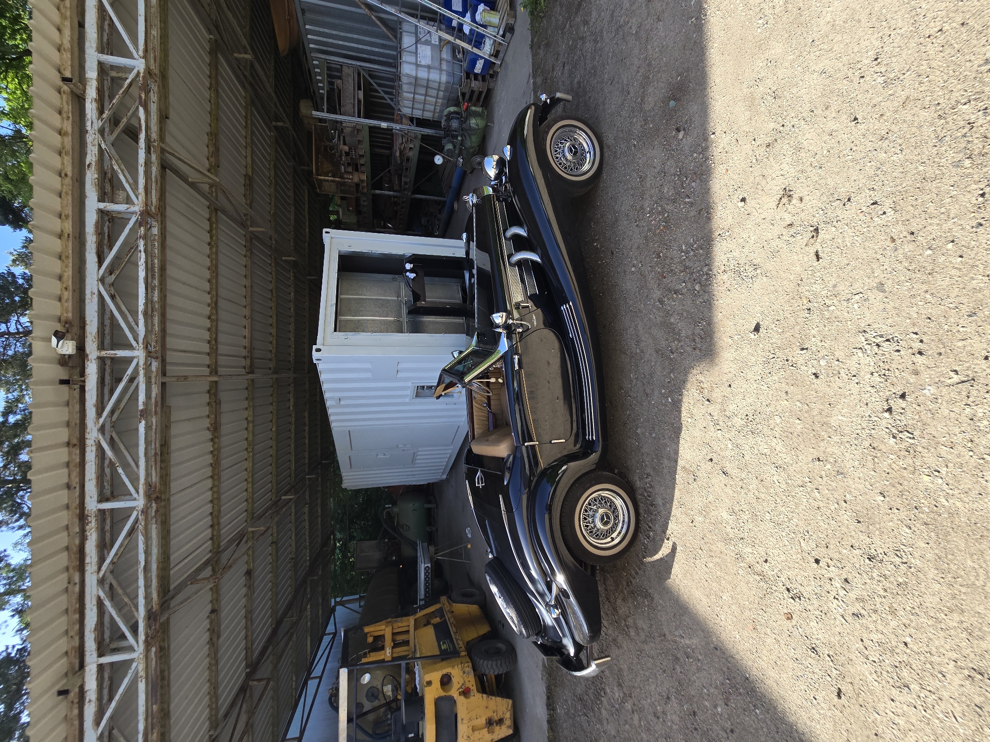 The image shows a vintage black car parked on a concrete surface next to an open-sided metal structure. Behind the car is a white shipping container. Various items, including crates, machinery, and a yellow forklift, are visible inside the structure. The location is an unidentified outdoor space.