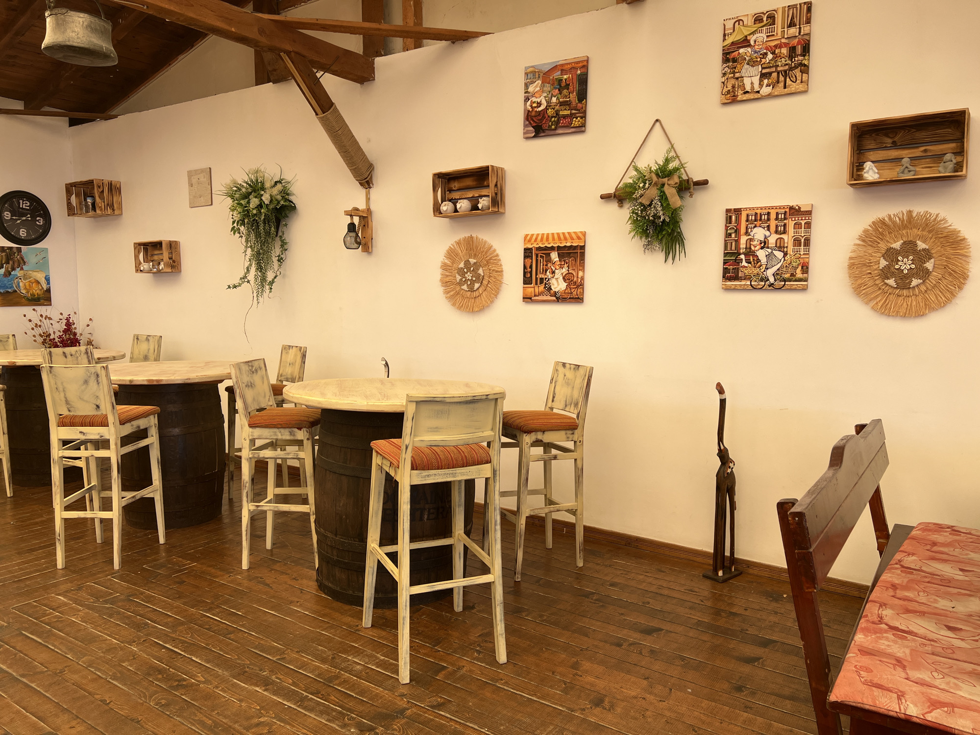 The image shows the interior of a restaurant located in Nesebar, Bulgaria. The restaurant has a rustic, country-style decor. The walls are painted white, and the ceiling is supported by exposed wooden beams. The floor is made of dark, polished wooden planks arranged diagonally.
Several round tables with light-colored tops are supported by dark barrels, functioning as bases. These tables are accompanied by tall, white-painted bar stools with orange striped cushions.
The walls are adorned with various decorative items. On the left, a large black clock with Roman numerals is mounted on the wall, next to a painting of a beer mug and a boat. Above the tables, wooden crates are mounted on the wall, some containing decorative items like ceramic pots or small figurines. Hanging plants are also visible on the walls, adding a touch of greenery.
Further to the right, there are several square paintings depicting chefs and market scenes. Between these paintings, a triangular arrangement of greenery and burlap hangs from a wooden dowel. A straw-like circular wall decoration is also present.
In the background, near the right wall, a tall, slender wooden sculpture of an animal, possibly a giraffe, stands on a small white base. A dark wooden bench with an orange patterned cushion is partially visible in the foreground on the right side of the image. The lighting in the restaurant appears to be warm and inviting, likely from ambient sources not directly visible. The overall impression is a cozy and charming dining establishment.