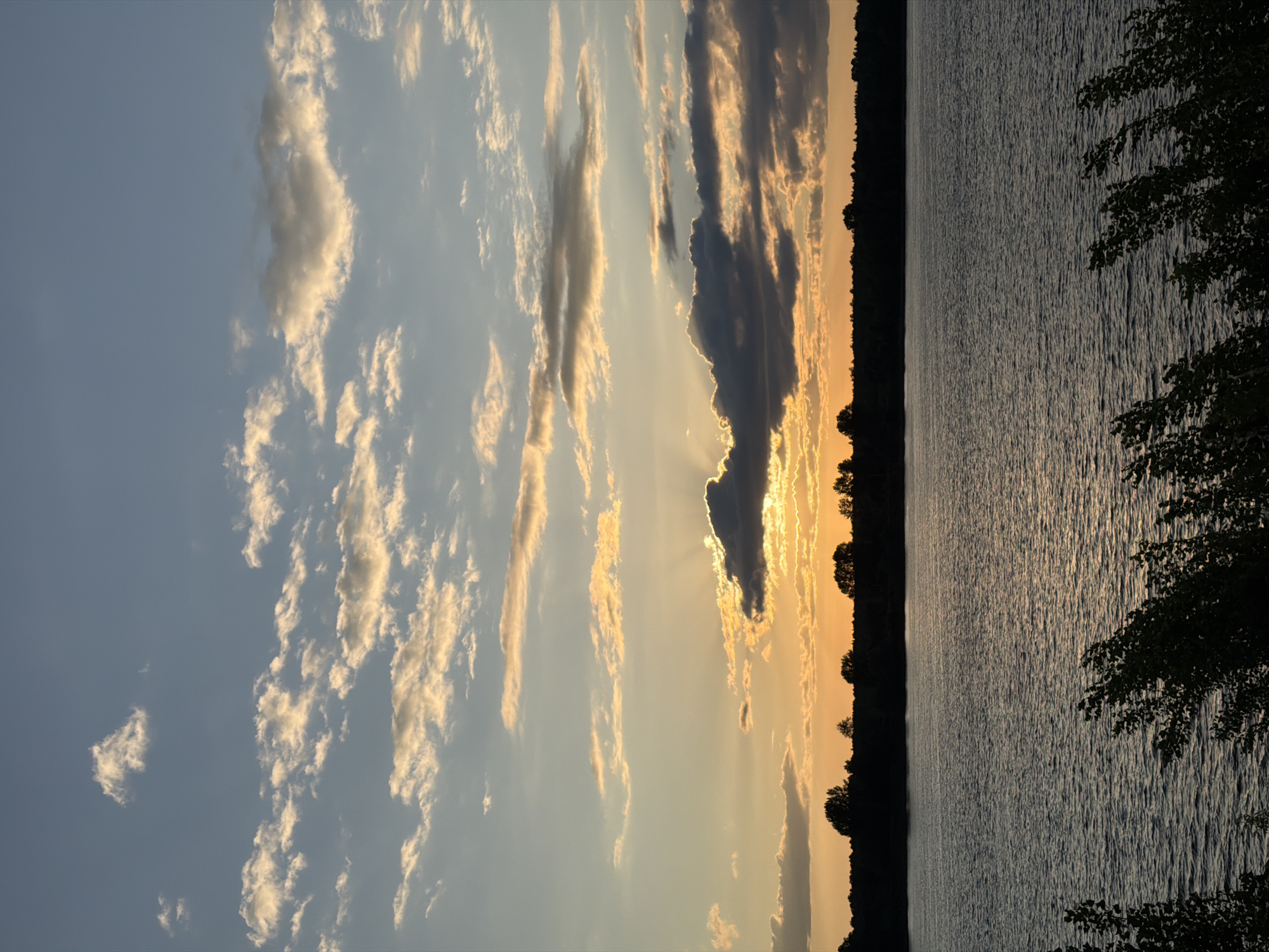 This image captures a serene sunset over the Kama River in Chaikovsky, Russia. The sky is a canvas of blues, grays, and oranges, with fluffy white clouds tinged with the golden hues of the setting sun. The sun's rays peek through a break in the clouds, casting a warm glow on the water. A dark silhouette of trees forms a line along the horizon, separating the sky from the tranquil river. The foreground is occupied by the dark, rippling surface of the Kama River, with hints of green foliage in the lower corners, suggesting the viewpoint is from the riverbank. The overall scene is one of natural beauty and peacefulness, characteristic of a calm evening in this Russian city.