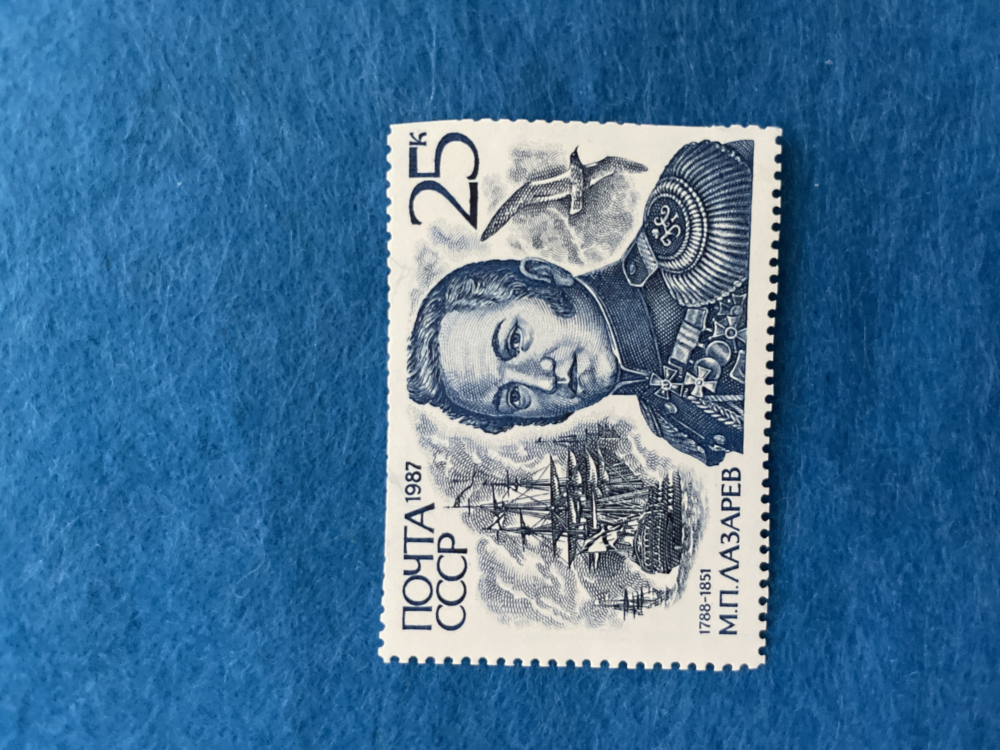 This is a close-up, overhead shot of a single postage stamp from the USSR, dated 1987, placed on a textured blue background. The stamp features a portrait of a man in military uniform, identified by the text "М.П. ЛАЗАРЕВ 1788-1851" and "ПОЧТА СССР". He wears a highly decorated jacket with medals and a distinctive hat. Above him, a seagull is depicted in flight, and below the portrait is a sailing ship. The stamp's denomination of "25K" is visible in the upper left corner. The overall tone is historical and commemorative. The background appears to be a fabric with a slightly fuzzy texture.