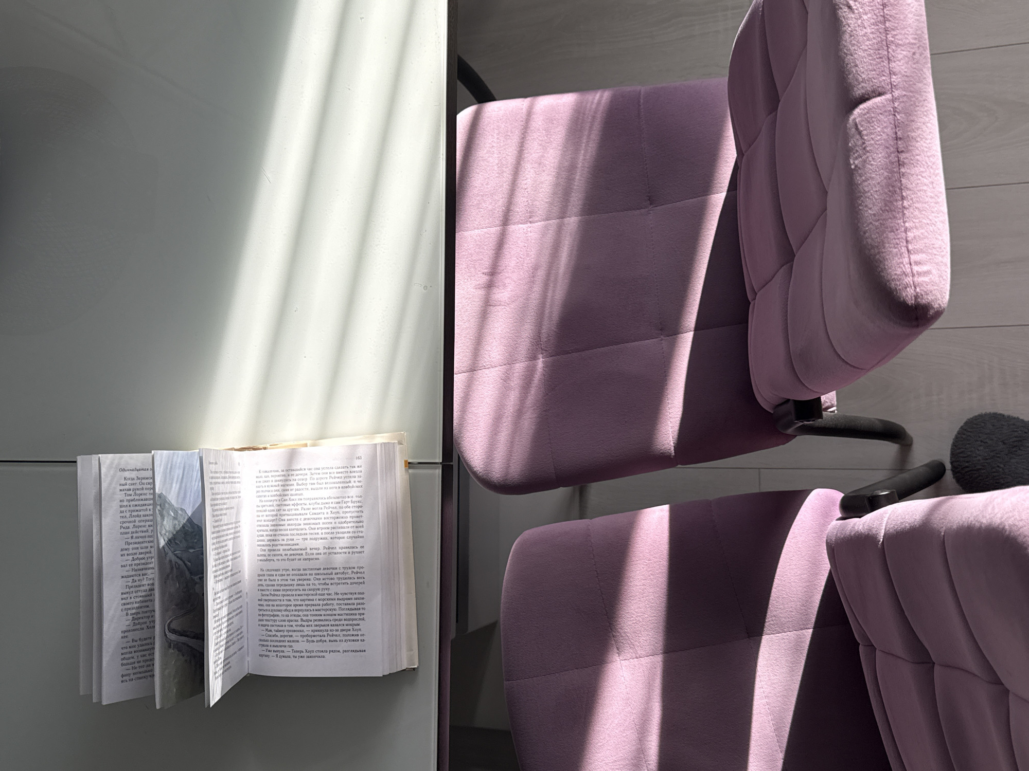 An open book lies on a white desk with diagonal shadows cast across it. Two upholstered chairs, one partially visible in the foreground and another to its right, are positioned in front of the desk. The chairs are a light purple color and have a tufted design. The room appears to be an indoor space with a wooden floor visible beneath the chairs. The lighting suggests it is daytime, with sunlight streaming in and creating the shadows. The overall scene is quiet and still, with no people present.