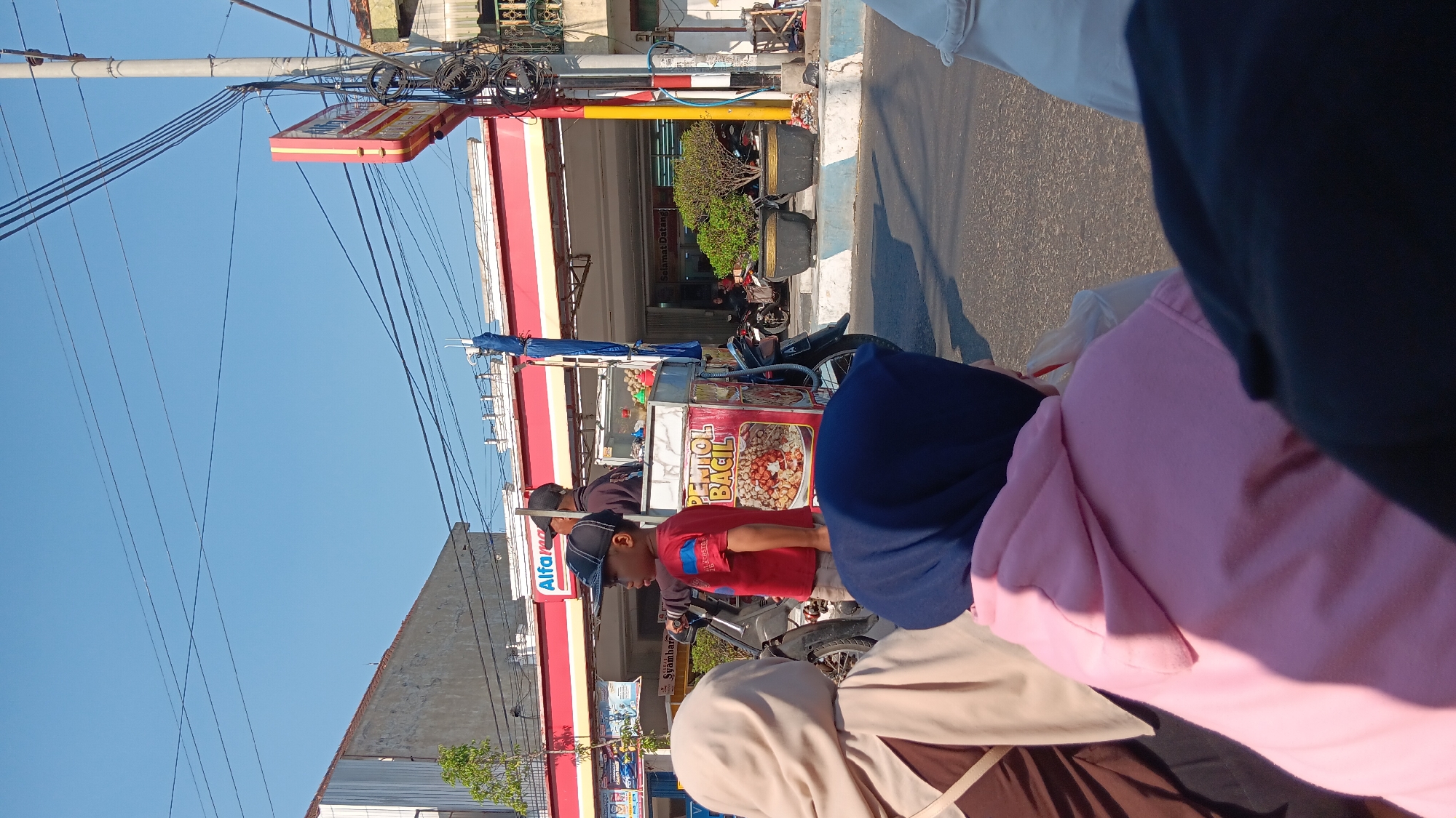 In Warujayeng, Indonesia, on a bright, sunny day, a young boy in a red t-shirt and black baseball cap stands near a food cart that is attached to a motorcycle. The food cart has a picture of a dish on it and the words "PENTOL BACIL". Behind him, a man in a dark shirt and black cap is on the motorcycle. Several people are visible in the foreground, one in a pink hoodie and another in a light brown headscarf, obscuring most of their faces. They appear to be a group of friends or family. In the background, there is a building with a red and white facade that has "ALFAMART" signage, indicating a convenience store. The street is paved, and there are power lines and a traffic light pole visible. The overall scene depicts a typical street view with a food vendor and shoppers in a busy Indonesian town.