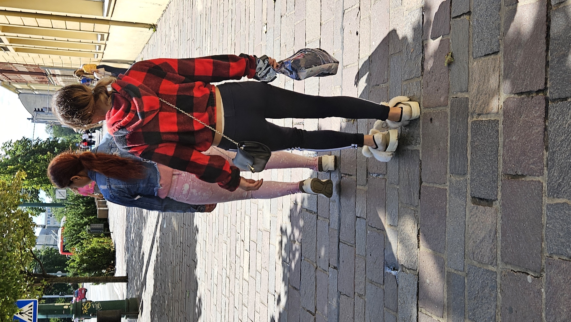 The image shows two young females walking on a cobblestone street in Prešov, Slovakia, on a sunny day. The female in the foreground, on the right, is wearing a red and black checkered hoodie, black leggings with a white stripe, and white fluffy slide sandals. She is carrying a dark grey plastic bag in her right hand and has a black purse with a gold clasp slung over her left shoulder. The female in the background, on the left, is wearing a denim jacket over a pink top and light pink patterned leggings. She has her red hair in a ponytail. The street is paved with irregular cobblestones, and shadows from trees and buildings are cast across the ground. In the background, there are buildings with yellow facades and windows, and some greenery. Other pedestrians are visible further down the street.