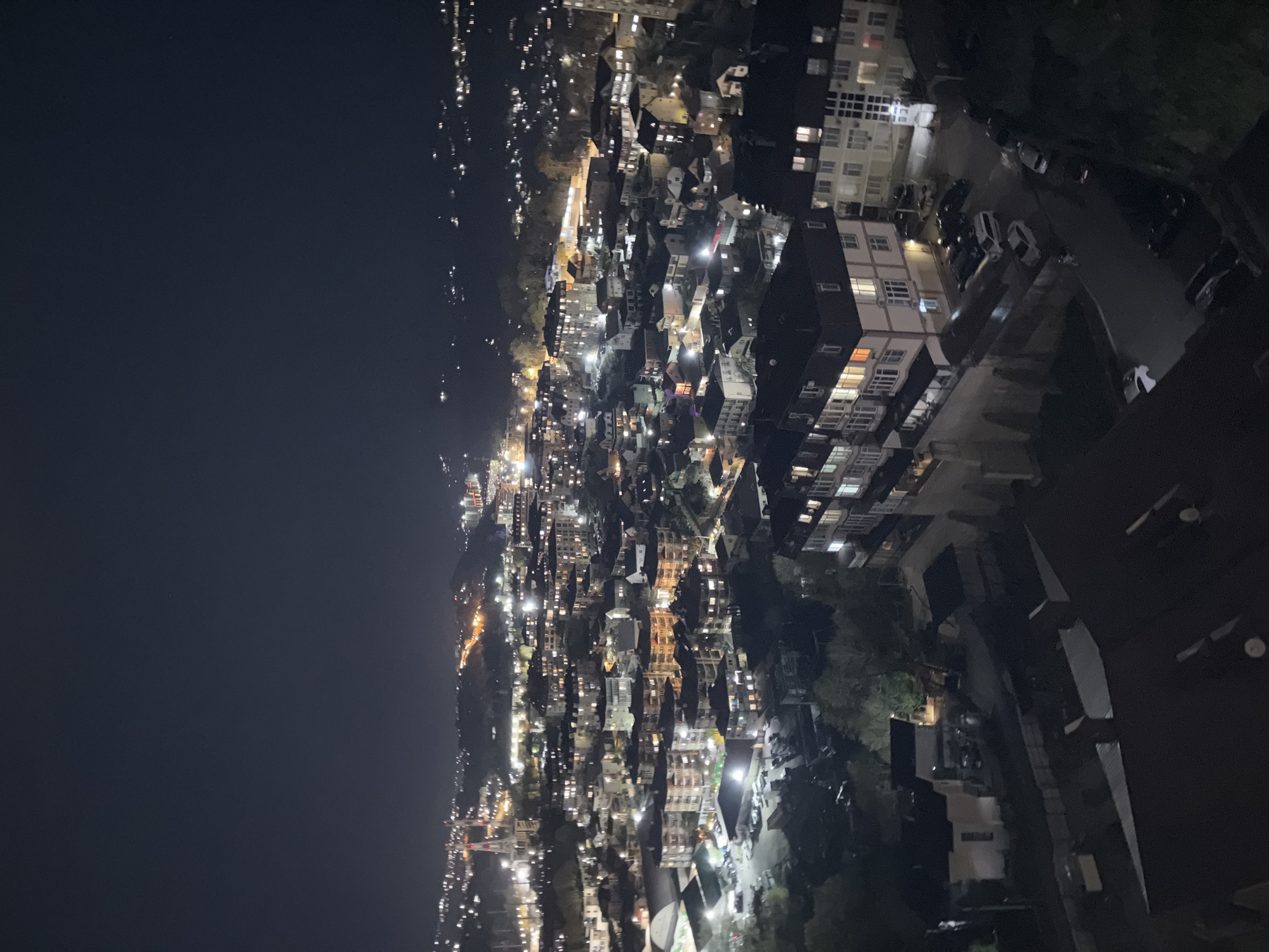 An aerial shot shows a brightly lit city at night. The city has a high density of buildings and the lights from cars and buildings create a stark contrast against the dark sky. A large dark body of water, likely the Black Sea, is visible on the left side. The city is likely Sochi, Russia, due to its coastal location and dense urban environment.