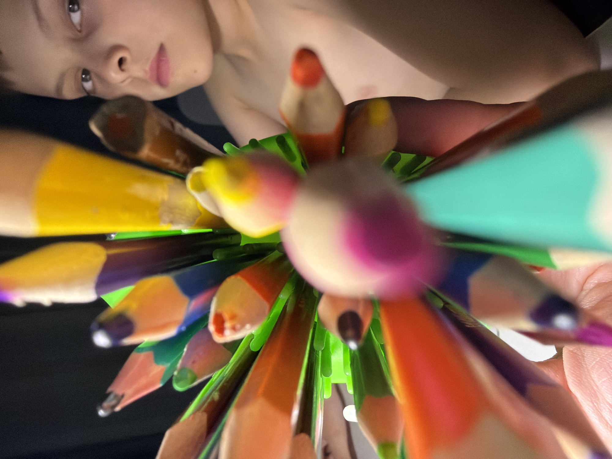 The image depicts a close-up, low-angle view of a collection of colored pencils held in a green plastic pencil holder. The pencil tips are in focus, creating a radial pattern. At the top of the frame, a child's face is visible, appearing to look down at the pencils. The background is blurred. The location of the image in Votkinsk, Russia is not discernible from the visual information provided.