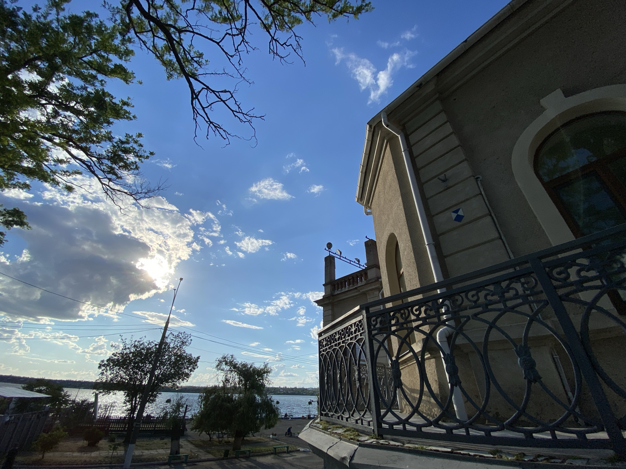 The original image is a wide-angle shot of a building with ornate metalwork on a balcony, taken from a low angle looking up towards the sky. The building is tan in color, with decorative stonework and a large arched window. A dark, wrought-iron balcony railing with an intricate pattern of ovals and decorative elements is in the foreground. The sky is a vibrant blue with scattered white clouds, and the sun is partially obscured by a large cloud formation on the left side of the frame, creating a bright lens flare effect. Tree branches with green leaves extend into the upper left corner of the image. In the background, a body of water, possibly a river or lake, is visible, with trees and some distant structures along the shoreline. A lamppost stands near the water's edge, and a few small figures are visible on the shore. The scene appears to be outdoors, with a pleasant, sunny day.