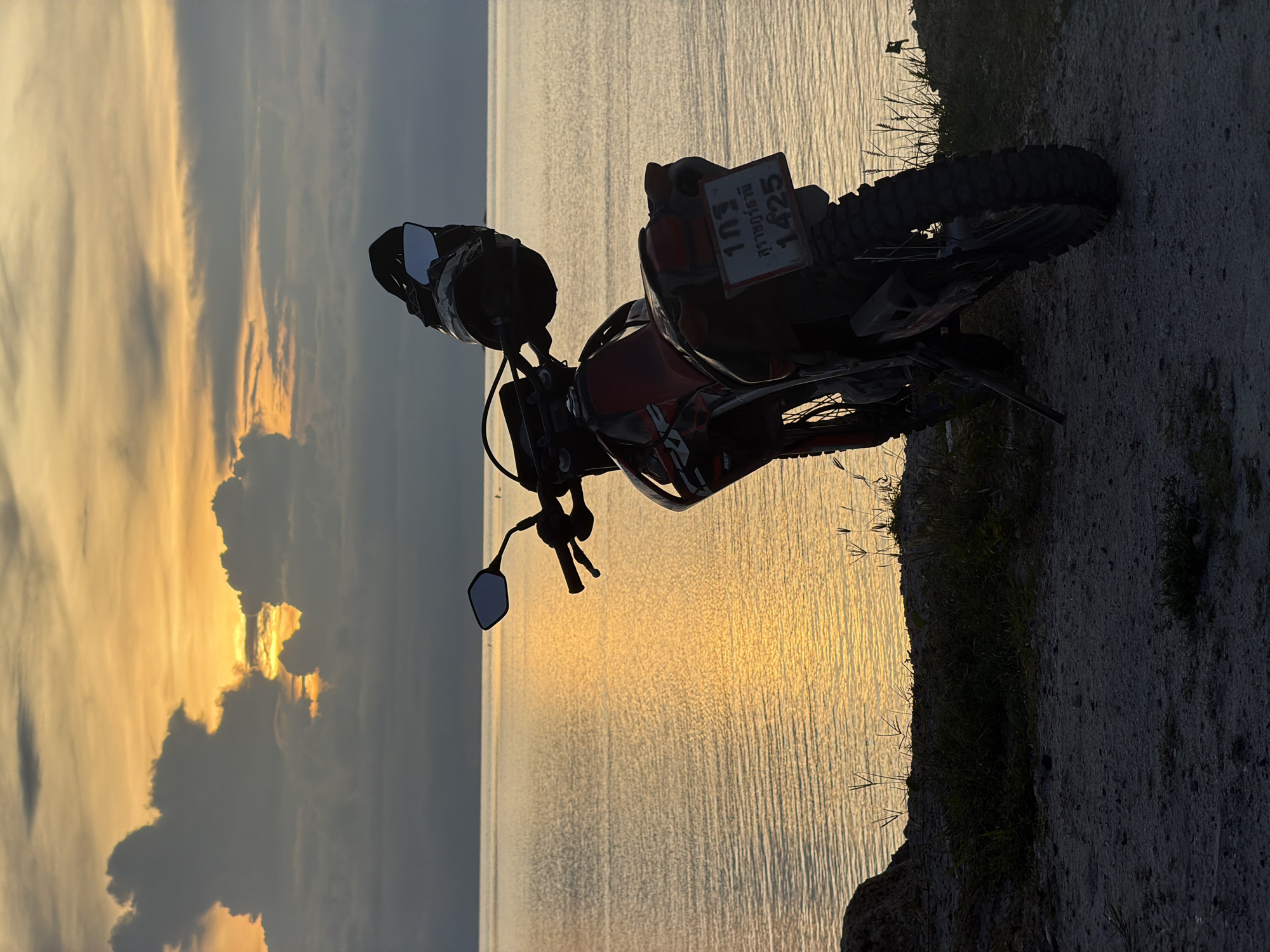 An outdoor, eye-level shot captures a dirt bike parked on a dirt road overlooking the ocean at sunset. The sky is a dramatic display of orange and grey clouds, with the sun peeking through them. The ocean is a shimmering expanse of gold, reflecting the sunset. The dirt bike is red and black, with a helmet perched on its handlebars. A license plate with Thai writing is visible on the back of the bike. The foreground shows a patch of dry grass and the dirt road. The scene is located in Tambon Ko Phangan, Thailand.