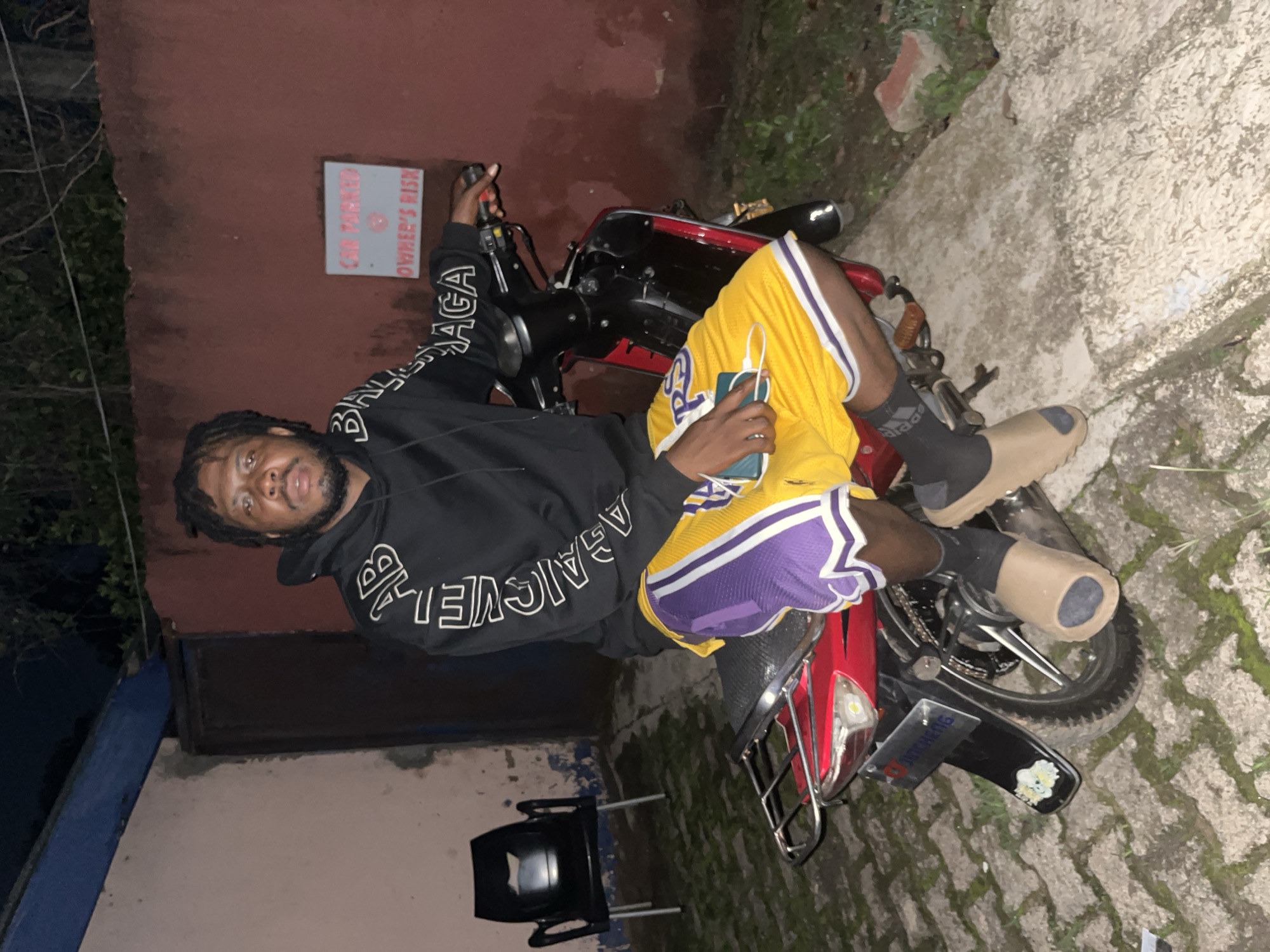 A man with dreadlocks is sitting on a red motorcycle at night. He is wearing a black hoodie with white text on the sleeves and yellow and purple basketball shorts. His feet are resting on the ground, and he is holding a cell phone in his left hand. In the background, there is a red wall with a sign that reads "CAR PARKED OWNER'S RISK". There is also a black plastic chair and some vegetation. The ground is paved with interlocking bricks. The lighting suggests that the image was taken in an outdoor urban environment, possibly a residential area or a commercial space, consistent with areas found in Jos, Nigeria.