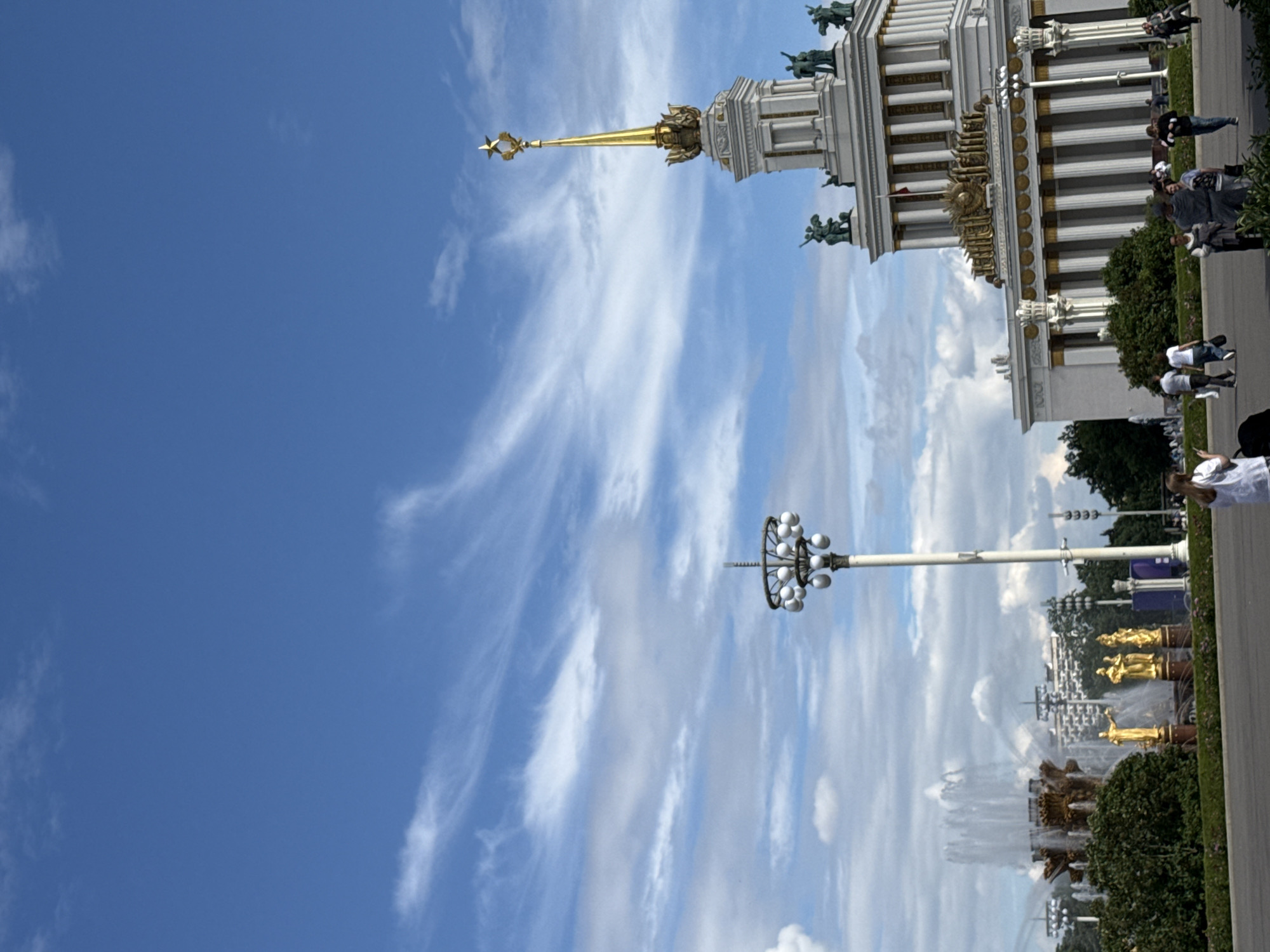 The image depicts a sunny day at a park in Moscow, Russia, with a clear blue sky dotted with wispy white clouds. The scene is dominated by a grand, ornate building with a golden spire reaching towards the sky. The building's architecture is elaborate, featuring columns and decorative elements. Several people are visible near the building, some walking and others standing, dressed in casual attire.
In the foreground, a tall lamppost with a circular arrangement of light fixtures stands out. Further into the background, a large, elaborate fountain is in operation, with water cascading down its tiers. Golden statues are prominently displayed on top of the fountain structure. The overall atmosphere appears lively and festive, suggesting a public gathering or tourist attraction. The presence of lush green trees and manicured hedges adds to the park-like setting. The time of day is likely midday, given the bright sunlight and clear sky. There is no visible text in the image.