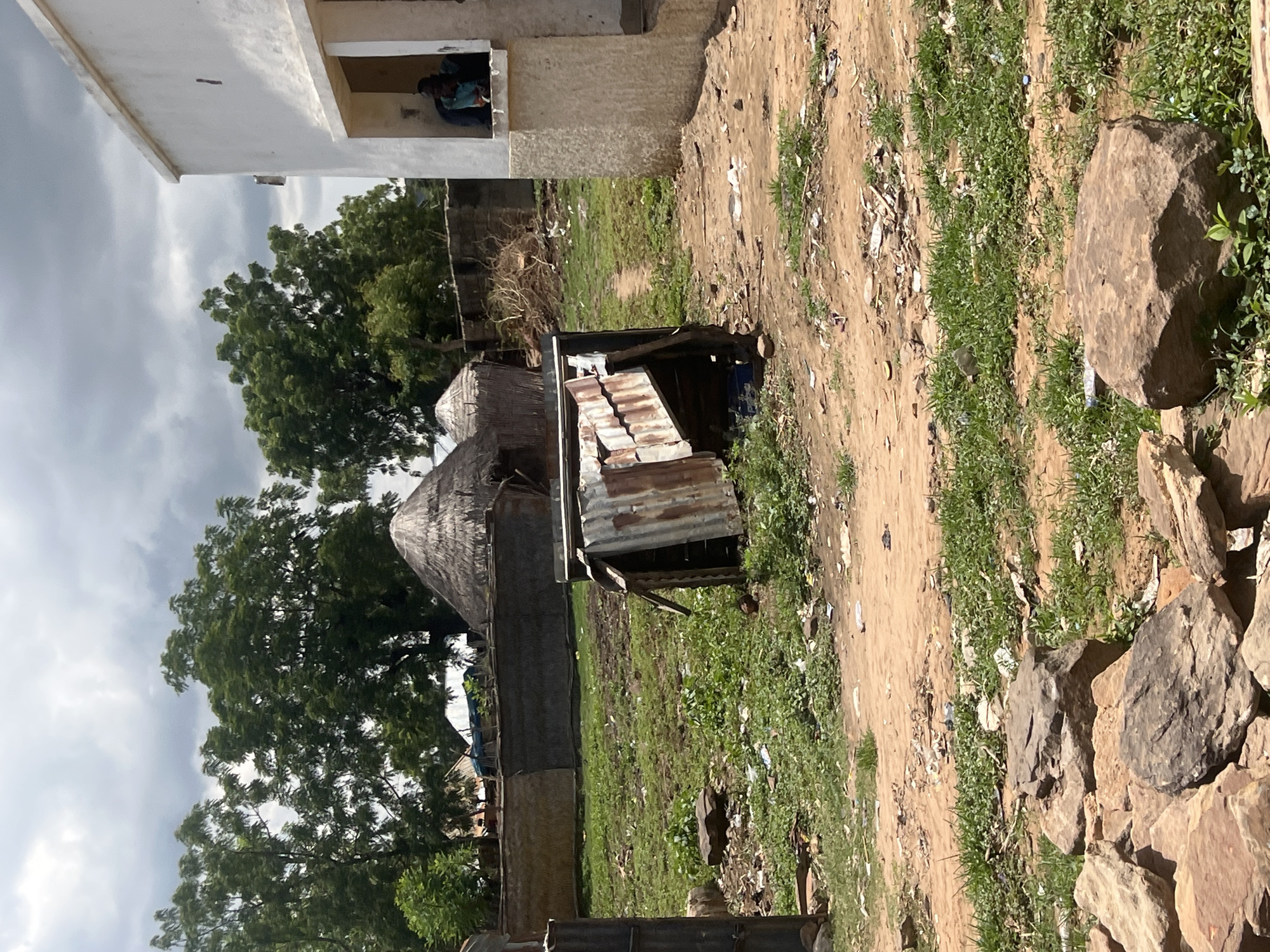 This image captures an outdoor scene in Sabon Layi Jessu, Nigeria, under an overcast sky. In the foreground, a collection of large, irregular rocks is arranged on a dirt and sparse grass patch, which is littered with debris. A dirt path extends from the foreground into the middle ground, where it passes a dilapidated structure constructed from rusty corrugated metal sheets, leaning towards the left. Beyond this, the middle ground features patches of green grass and dirt, also interspersed with various litter.
In the background, a barrier or fence made of woven materials or thin slats extends horizontally, separating the immediate area from more distant structures. Behind this fence and partially obscured by dense green trees, several traditional round huts with conical thatched roofs are visible. Other structures, including parts of what appears to be a blue tarp or roof, are also present in the far background.
To the right side of the frame, a two-story or raised single-story white building with a flat roof stands prominently. An open recess, resembling a small porch or large window, is visible on its lower section, supported by a concrete column. Within this recess, a person, appearing to be an adult male, is seated, facing outwards. They are dressed in dark clothing with a lighter blue or green garment visible underneath. Their posture is relaxed, and their gaze is directed towards the front, but no specific action is discernible beyond sitting.