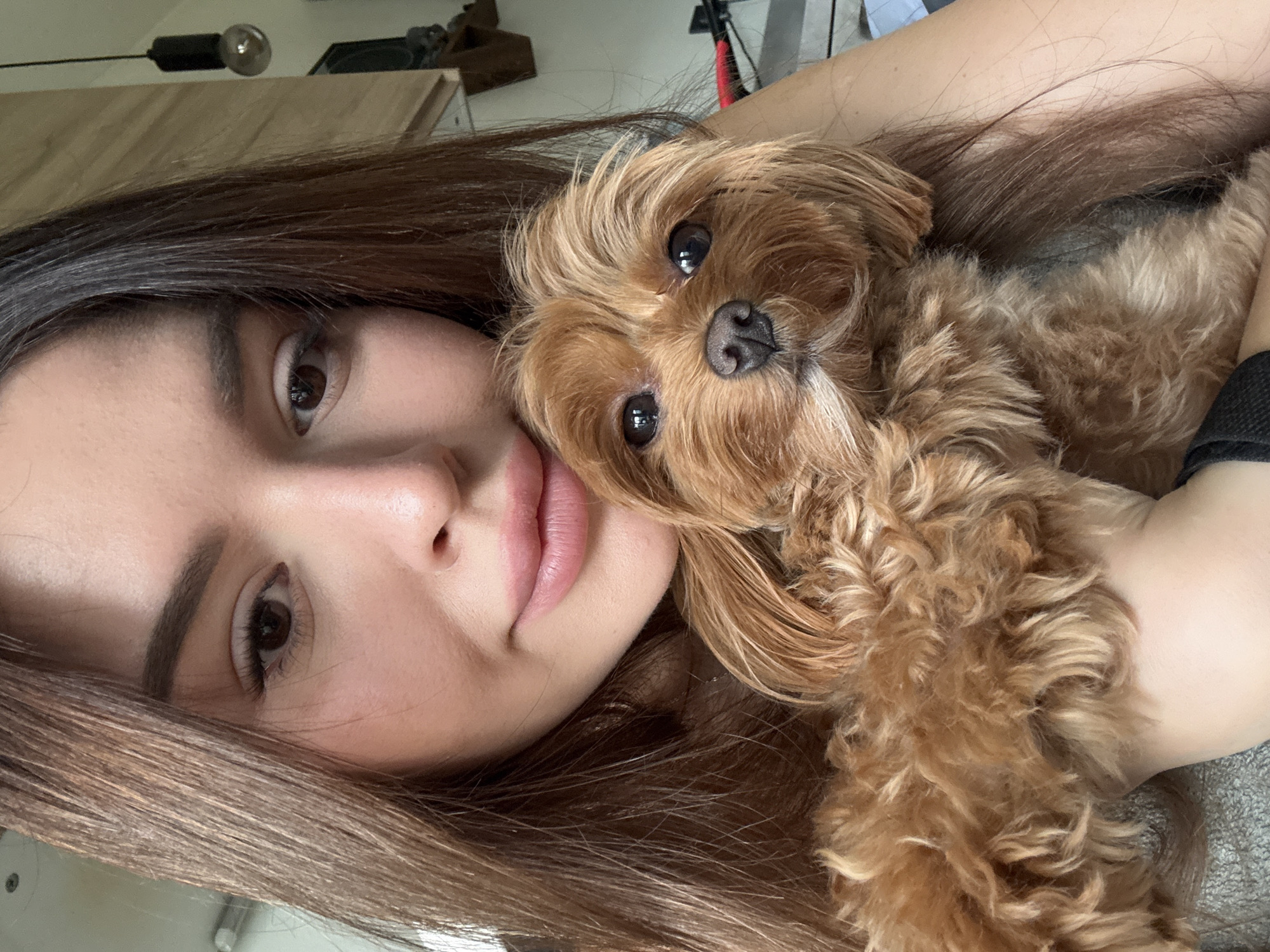 A close-up, selfie-style portrait shows a young woman with long, brown hair holding a small, tan dog. The woman's face is partially obscured by her hair and the dog. She has brown eyes and pink lips. The dog has dark eyes and a wet nose, with fluffy, reddish-brown fur. They appear to be indoors, with a wooden wall and a light fixture visible in the background. The image does not provide any information about the location being Prague, Czechia.