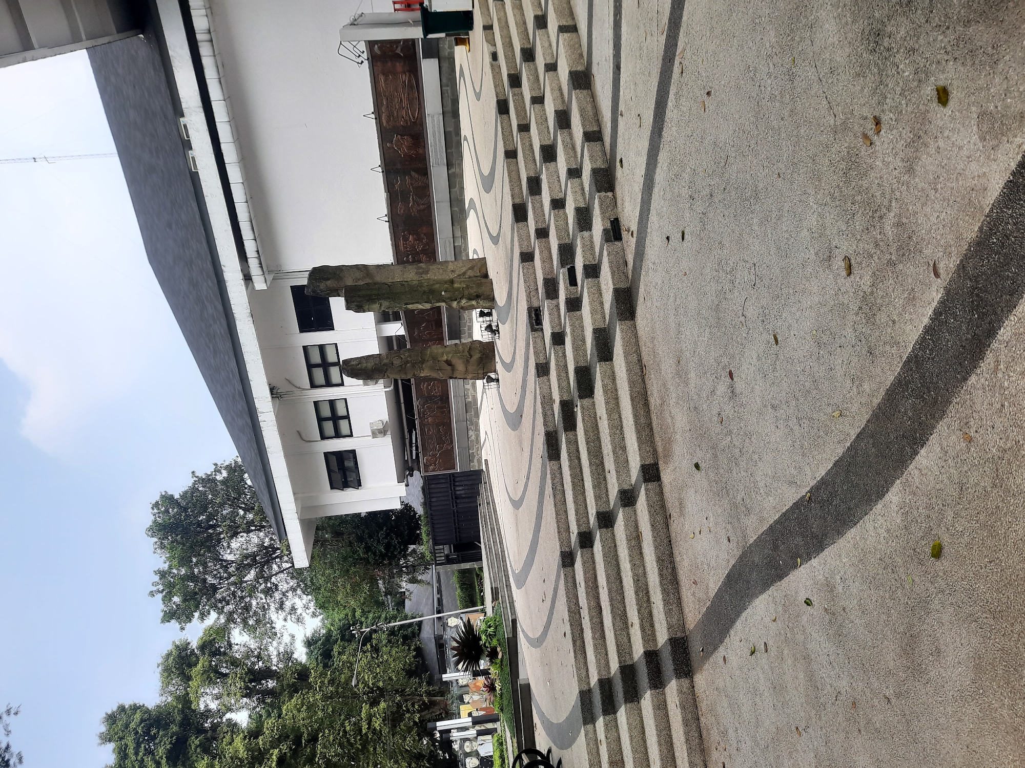 The image depicts an outdoor scene in Bandung, Indonesia, likely a public space or monument. In the foreground, a wide, multi-tiered set of concrete steps with dark grey accents creates a geometric pattern leading upwards. The steps are textured with small pebbles. The ground around the steps features curved, dark grey lines set against a lighter, pebbled concrete surface, with scattered fallen leaves.
Behind the steps, three tall, rough-textured stone pillars stand in a row. Behind the pillars, a white building with a dark, sloped roof dominates the background. The building has several dark-framed windows and visible air conditioning units. A long, dark bronze-colored relief mural depicting figures and scenes is mounted on the lower part of the building's exterior wall. To the left of the building, a black metal gate is visible, and beyond that, a collection of trees with lush green foliage.
To the far left of the image, an area with more detailed imagery is partially visible, including what appears to be a mural of several figures in uniforms and a small garden with a spiky plant. The sky above is overcast with a pale blue hue. The overall impression is of a somewhat monumental or commemorative outdoor area in a city setting, with a blend of modern architecture and natural elements.
