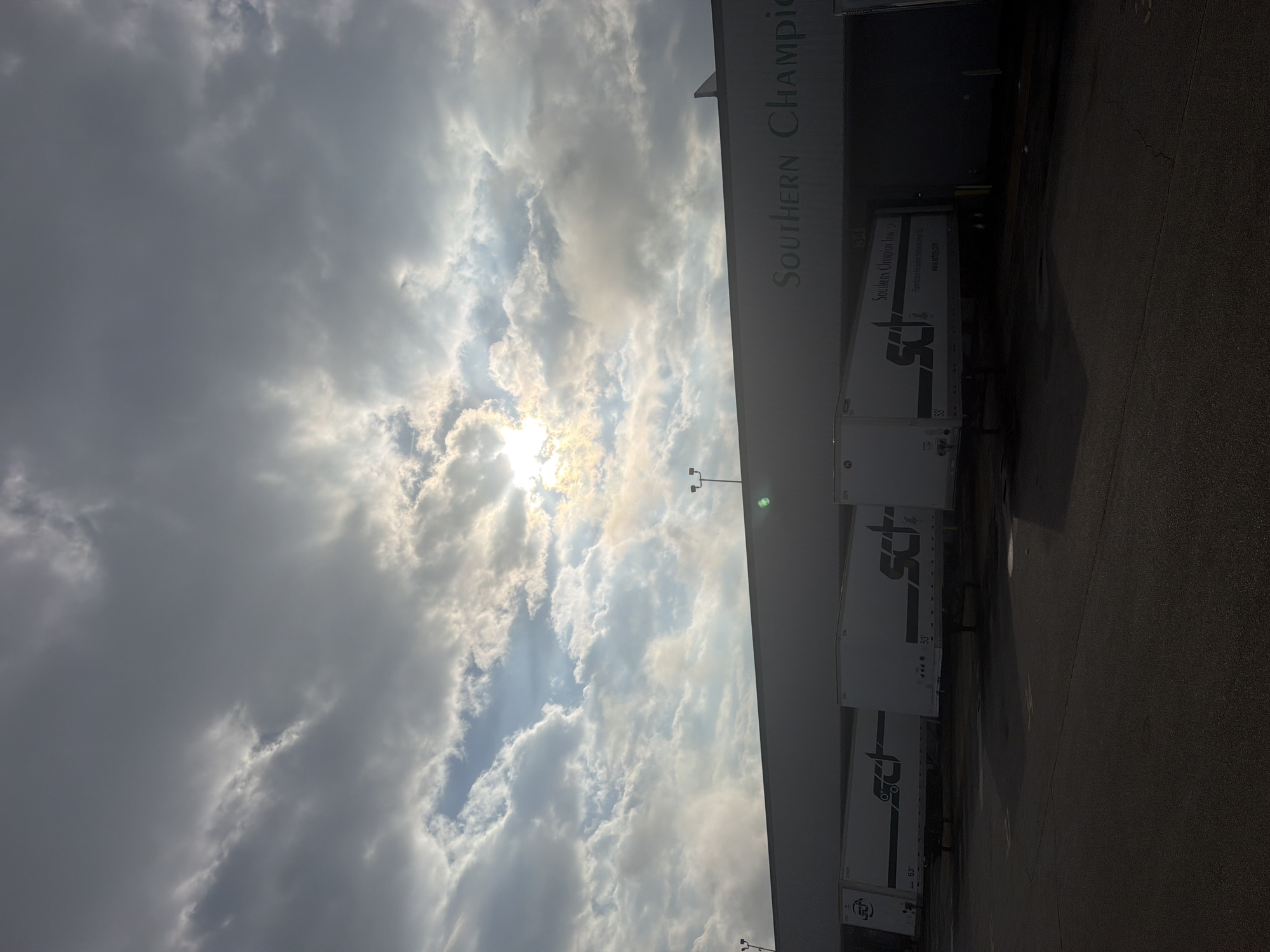 The image captures a low-angle view of a commercial building facade with the name "SOUTHERN CHAMPION" visible in green lettering across the top. Several white semi-trailer trucks are parked in front of the building, some bearing the "SCT" logo. The sky above is filled with dramatic clouds, with the sun partially obscured and shining through, creating a bright effect. The scene appears to be an industrial or logistics area, possibly a distribution center or warehouse. The ground in front of the building is paved, and streetlights are visible on poles. The overall lighting suggests it could be early morning or late afternoon, given the low sun angle and the contrast between the bright sky and the darker foreground.