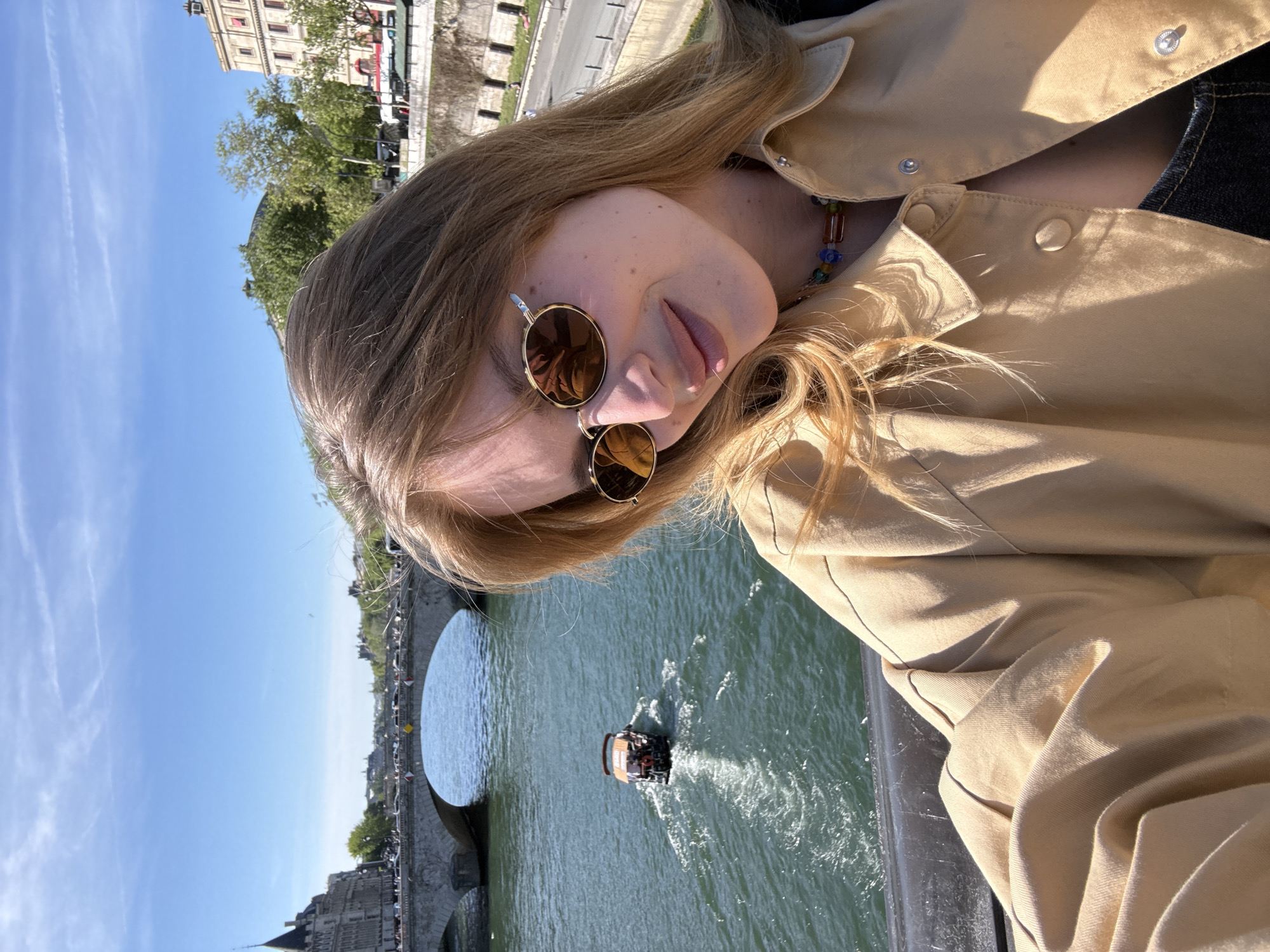 The image, oriented at a tilt, captures a close-up of a person, likely a young woman, on a bright, sunny day in Paris, France. She has long, light brown or blonde hair and fair skin with visible freckles. Her eyes are obscured by round, gold-rimmed, reflective sunglasses. Her lips are slightly parted, and she appears to be looking towards the camera.
She is wearing a tan or beige collared jacket with snap buttons, and a multicolored beaded necklace is visible around her neck, peeking out from under the collar. A dark garment, possibly denim, is also visible under her jacket.
In the background, under a clear blue sky with faint contrails, a historic stone bridge with multiple arches spans a river. A small motorized boat is visible on the river, leaving a white wake behind it. Along the riverbanks, buildings and trees can be seen, characteristic of an urban European setting. The overall atmosphere suggests a leisurely daytime moment, possibly a selfie taken while sightseeing. There is no visible text in the image.