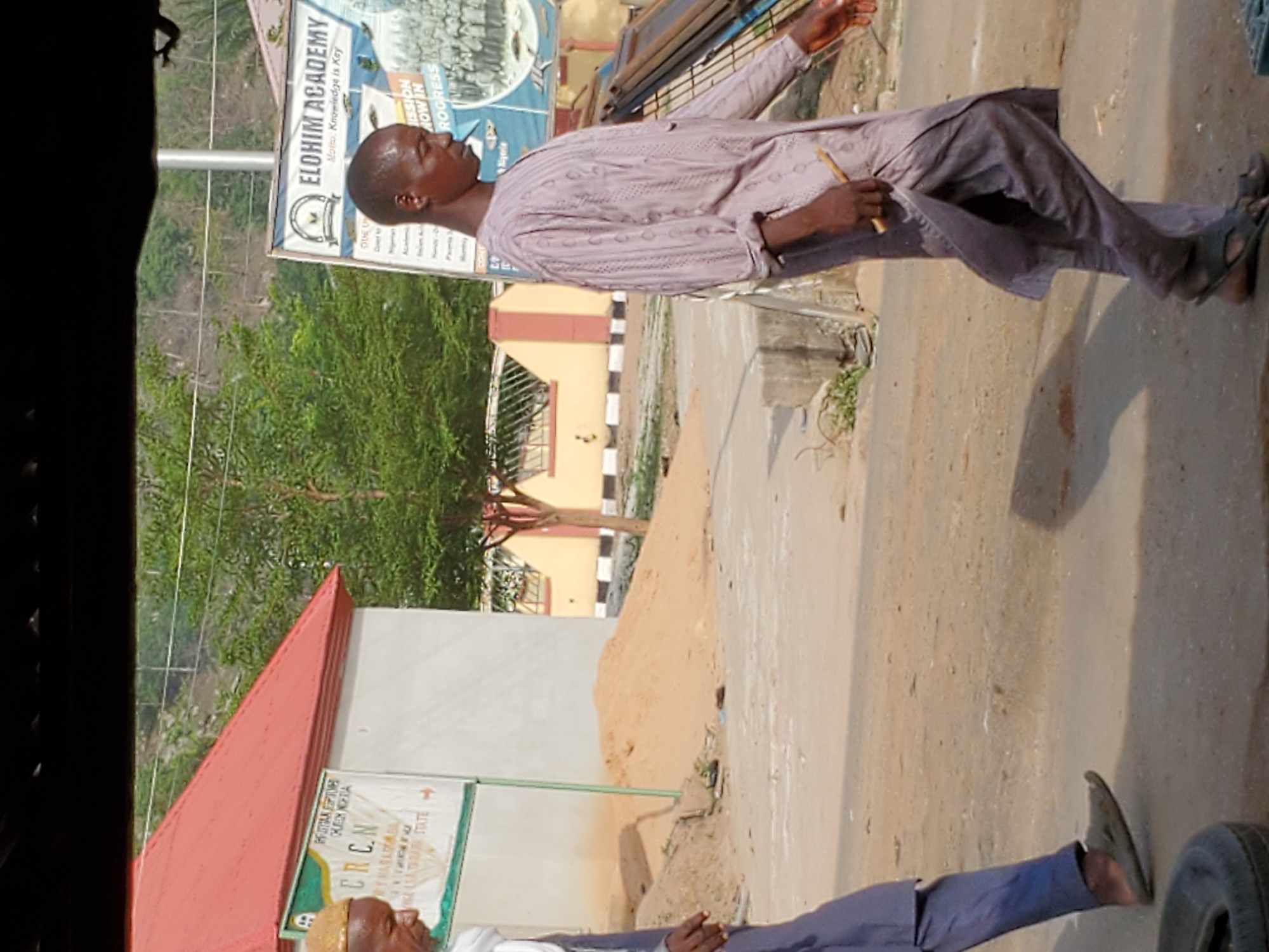 This media file captures an outdoor scene in Mararaba, Nigeria, during daylight hours with bright sunshine, indicated by strong shadows. Two men are visible in the frame.
In the midground, a man with a shaved head stands, facing towards the left side of the image. He is dressed in a light-colored, patterned, traditional long-sleeved tunic (possibly an agbada or kaftan) over dark trousers, paired with dark sandals. He is holding a slender, light-colored object, which could be a chewing stick or a pen, in his right hand, while his left hand is slightly raised.
In the foreground, another man is partially visible, mainly his lower body, wearing dark blue trousers and dark shoes/sandals. A car tire is also partially visible on the ground in the bottom right corner.
The background features several elements: a road or dirt path, lush green trees and vegetation, and various buildings. One building in the lower left has a distinctive red corrugated roof. Another building in the distance is light yellow with black and white striped roadside curbs. A tall white pole stands vertically near the center.
Several signs are present in the image. A prominent white and blue sign for "ELOHIM ACADEMY" is visible behind the man in the midground, with the motto "Knowledge is Kay" (likely "Key") and partial text reading "MISSION NOW IN PROGRESS". On the building with the red roof, a green and white sign displays "CRCN" and "GATARI STATE", along with other less clear text like "OUEU" and "Dover k".