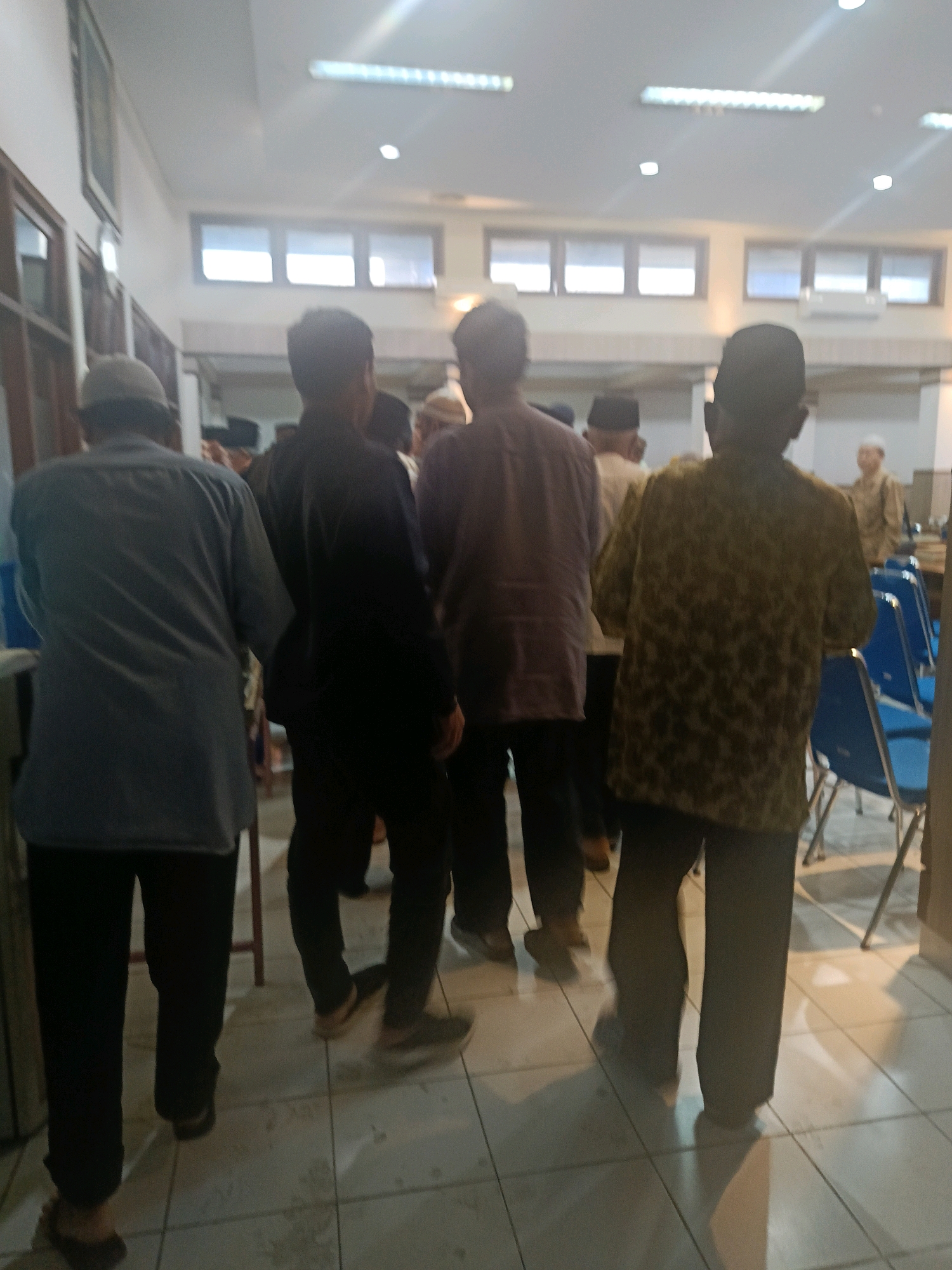 The image captures a group of men, likely in a community hall or indoor gathering space, in Yogyakarta, Indonesia. Several men are seen from behind, facing away from the camera. The man in the foreground to the left wears a grey long-sleeved shirt and dark pants, with a light-colored cap. To his right, another man is dressed in a black shirt and pants. The man in the center, also viewed from the back, is wearing a dark patterned long-sleeved shirt and dark pants. Further to the right, a man in a green and brown patterned shirt and dark pants stands with his back to the camera. In the background, other individuals are partially visible, including one man in a light-colored shirt and cap. The environment appears to be an indoor space with tiled flooring and large windows with blinds in the background. Fluorescent lighting fixtures are visible on the ceiling. Blue chairs are arranged in rows to the right of the frame. The overall impression is that of a gathering or event taking place.