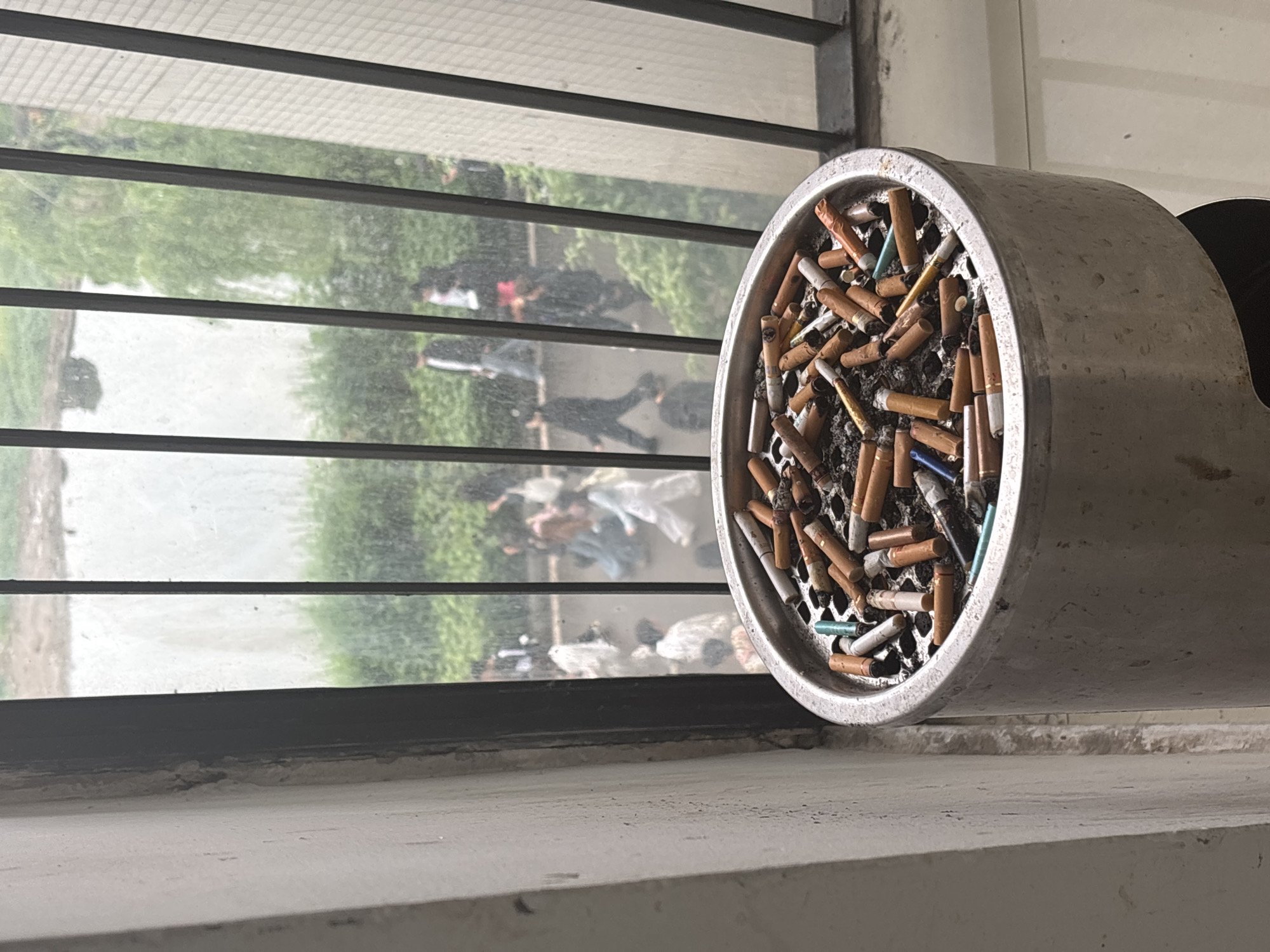 An eye-level, indoor shot shows a metal ashtray filled with cigarette butts and ash positioned on a white windowsill, against a black-framed window with horizontal metal bars. Reflected through the window, a scene is visible outside, showing several people walking on a concrete surface, along a green, tree-lined area. The scene appears to be in daylight. Based on geotags, the location is Jiangjin, China.