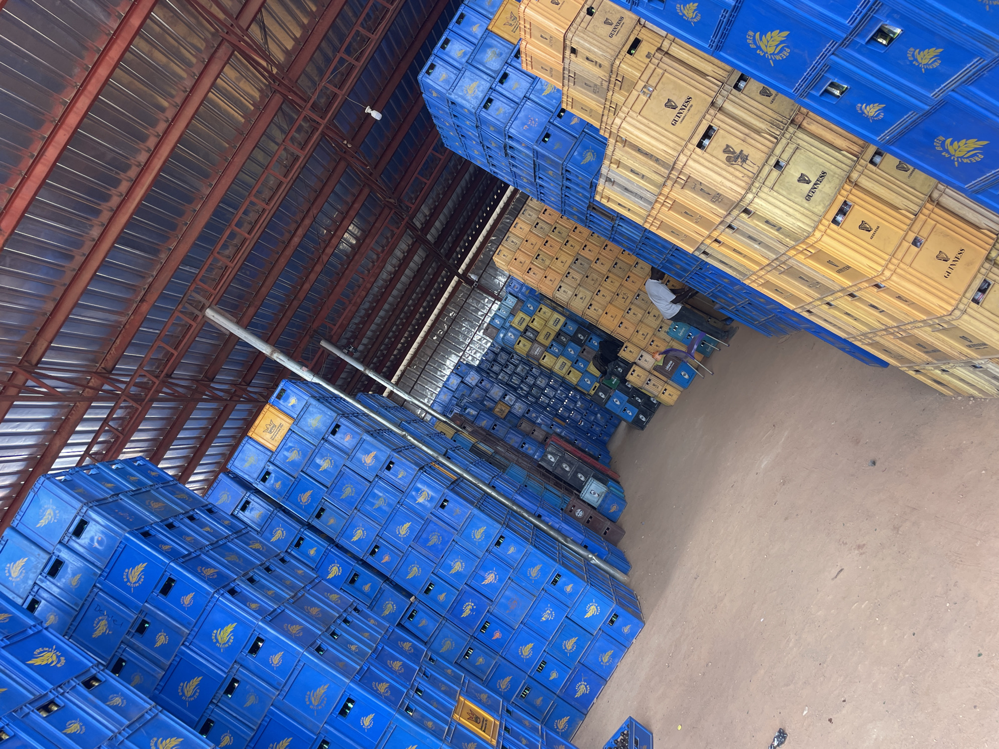 The image depicts a large storage warehouse in Gura Top, Nigeria. Stacks of blue and tan plastic crates, presumably for beverage bottles, dominate the scene, reaching high towards the metal roof of the warehouse. A single individual, wearing a light-colored shirt, is seated amongst the stacks of crates in the distance. The floor is bare earth. The lighting is a mix of natural light from the warehouse roof and artificial lighting.