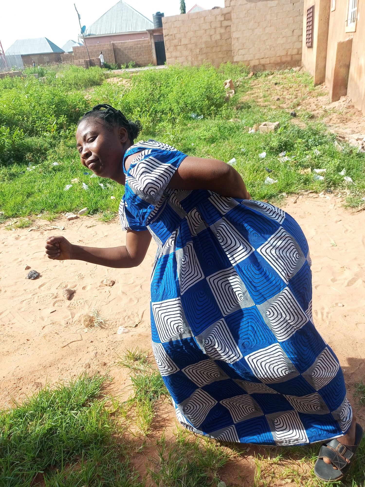 The image depicts a woman in a blue and white patterned dress, bending forward with her right arm extended and fist clenched. She is standing on a dirt path with some grass growing around her feet. In the background, there are buildings constructed with light-colored bricks, and some vegetation. The scene appears to be an outdoor area in a residential neighborhood. The location is identified as Tempure, Nigeria.