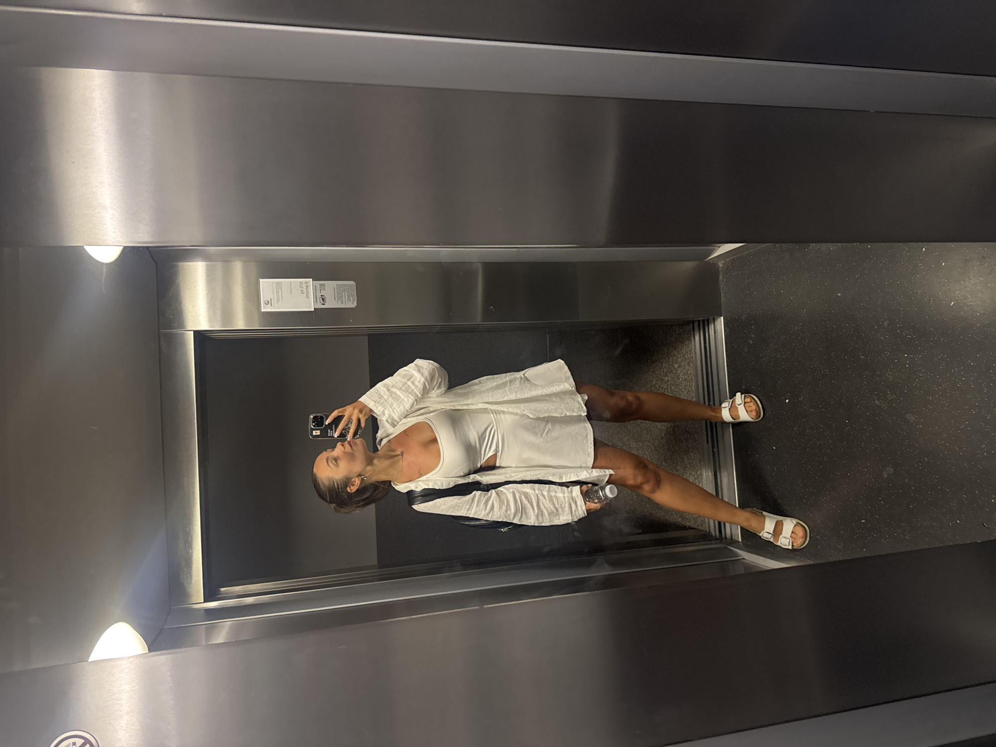 An outdoor shot, taken in the city of Budva, Montenegro, shows a young woman posing for a selfie in an elevator. The elevator's interior is made of stainless steel, with a light fixture visible on the ceiling. The woman is wearing a white cutout dress, a white linen shirt, and white sandals. She has a black backpack slung over her left shoulder and is holding a water bottle in her right hand. The elevator's floor is dark and appears to be made of speckled material. A sign with text and a diagram is affixed to the wall of the elevator.
