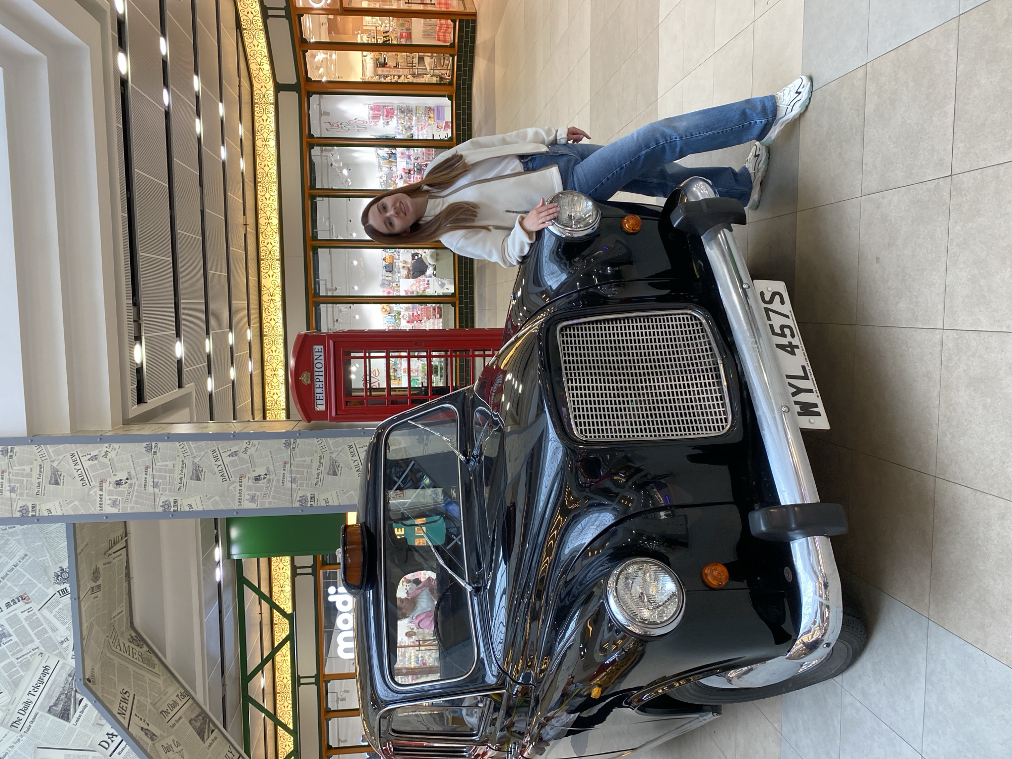 This is a vertical, full-body photograph of a young woman leaning on the hood of a black London taxi. She is wearing a white hooded jacket, blue jeans, and white sneakers. Her hair is long and light brown, and she has a slight smile on her face as she looks at the camera. The taxi is positioned in the foreground, with its distinctive grille and chrome bumper clearly visible.
Behind the taxi, a traditional red British telephone booth stands out. The background suggests an indoor setting, possibly a shopping mall or a themed exhibition, with display windows of shops visible. The ceiling has modern lighting fixtures, and the floor is tiled. The overall impression is a playful and slightly staged scene, bringing a piece of London to another location.
On the taxi, a license plate is visible with the text "WYL 457S". In the background, there's a sign with the word "modi" in large letters. There is also some text on what appear to be decorative newspaper clippings on a wall, including "The Daily Telegraph" and "London Bridge."
Given the context of Saint Petersburg, Russia, this scene likely represents a themed attraction or a display designed to evoke a British atmosphere. The lighting suggests it is daytime indoors. The woman's pose and smile convey a relaxed and happy mood.
