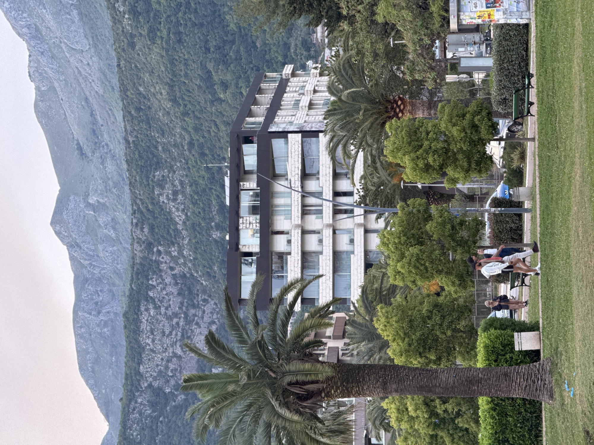 The image captures a scene in Budva, Montenegro, displaying a juxtaposition of urban architecture and natural landscapes. A modern multi-story building, constructed with alternating black and white horizontal sections, stands amidst lush greenery and palm trees. The building's design includes numerous windows, reflecting the daylight. To the left, a massive, rocky mountain dominates the horizon, its slopes covered in vegetation. A park-like area is visible in the foreground, with a patch of green grass, trees, and several people. A few individuals are standing and engaging in a conversation, while others are seated on a bench. The overall atmosphere suggests a harmonious blend of urban development and natural beauty, characteristic of Budva's coastal setting.