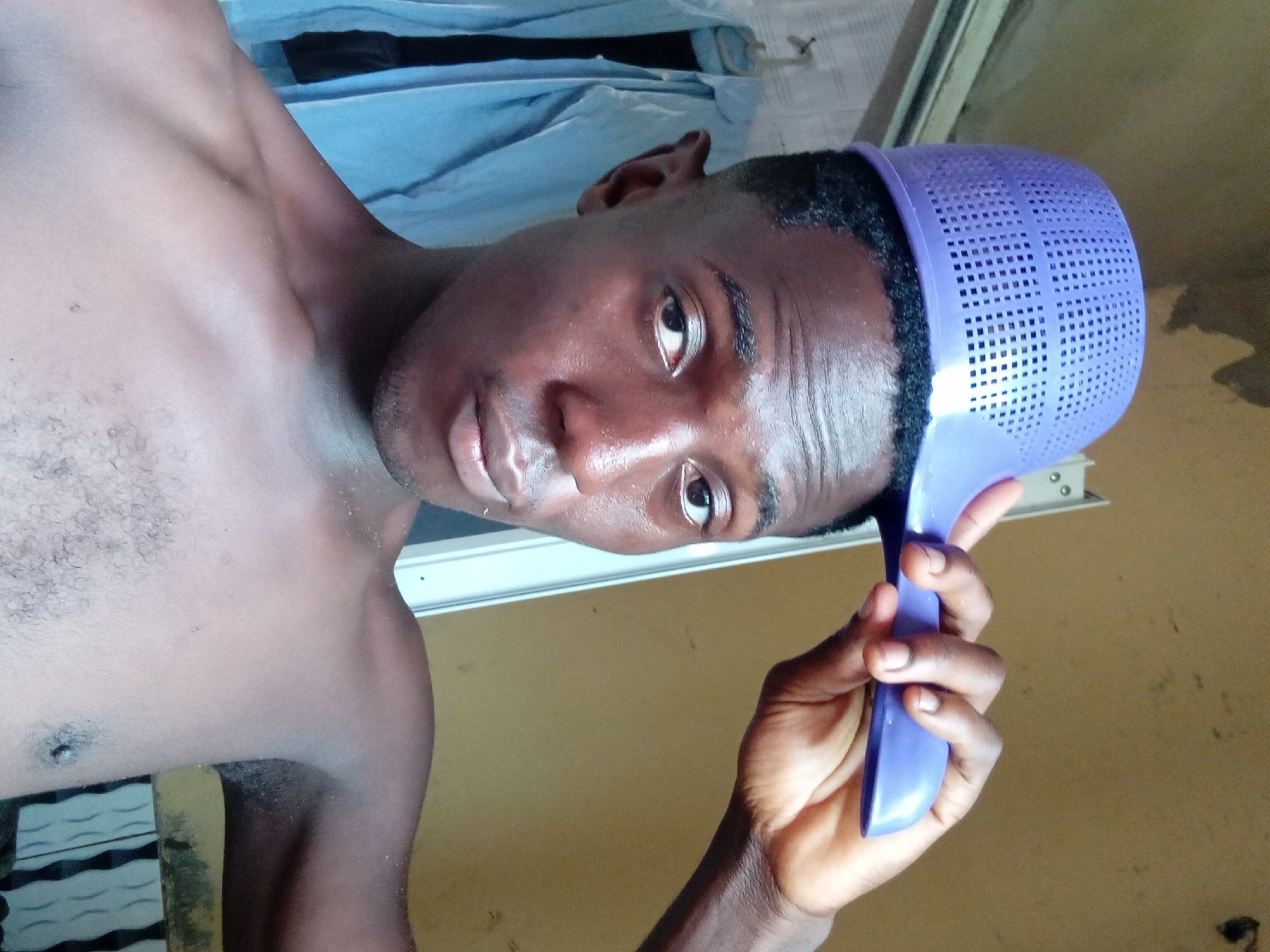 A shirtless man is holding a purple plastic colander on his head. He is looking towards the camera. The location is Akwete, Nigeria.