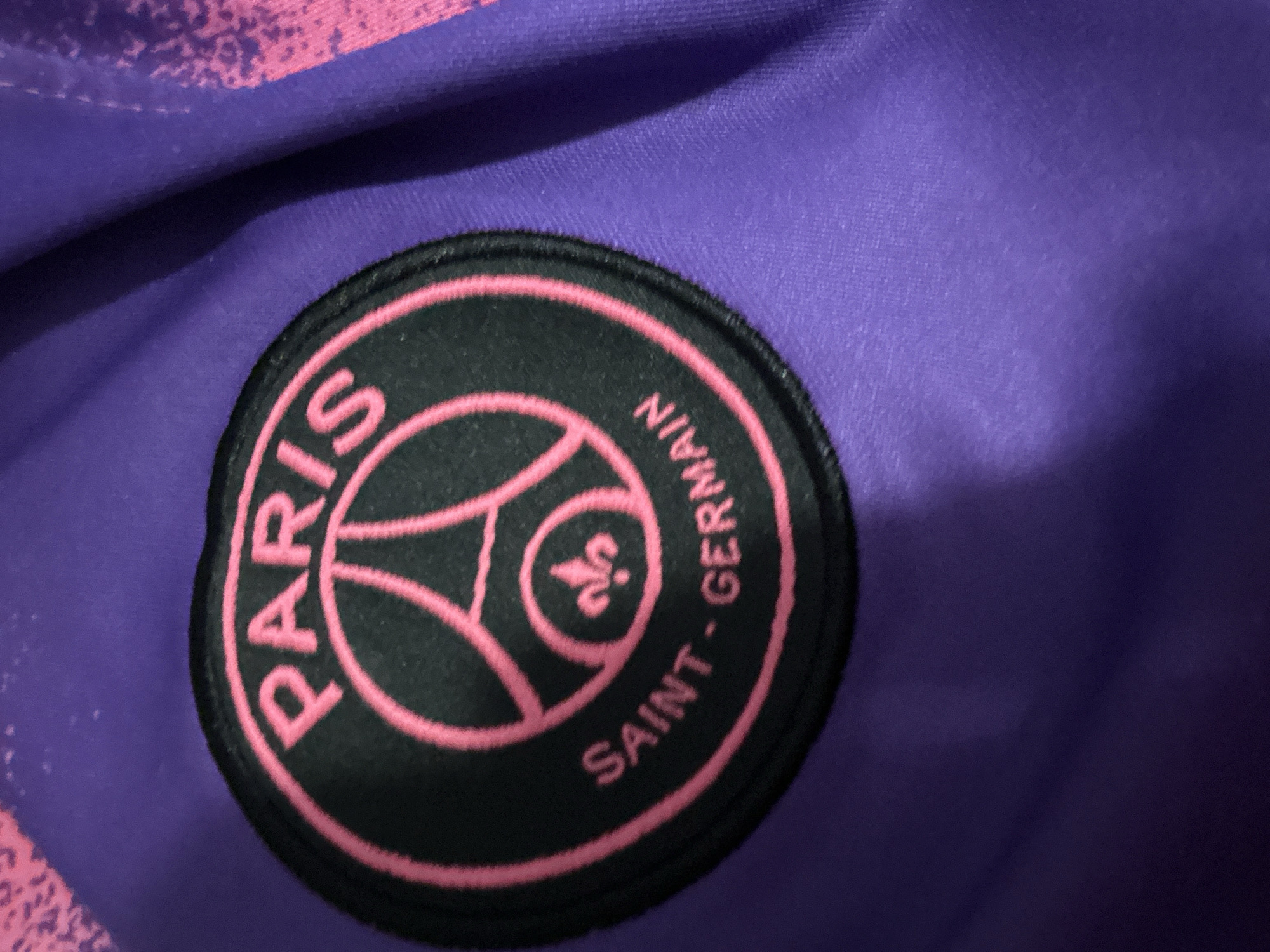 The image shows a close-up of a Paris Saint-Germain football club crest. The crest is circular with a black background and pink lettering and details. The word "PARIS" is written in pink capital letters at the top of the crest. Below that, there is a pink outline of a football stadium with a silhouette of the Eiffel Tower inside. The words "SAINT-GERMAIN" are written in pink capital letters along the bottom curve of the crest. The crest is embroidered onto a purple fabric, which appears to be part of a football jersey or other athletic apparel. The fabric has a subtle texture and a slight sheen. Some parts of the fabric are creased, indicating it is not perfectly flat. There are some lighter pink, abstract patterns visible on the upper right side of the purple fabric, possibly part of the garment's design. The lighting in the image is somewhat dim, creating shadows on the fabric and making the colors appear richer. The focus is sharp on the crest, while the surrounding fabric is slightly out of focus.