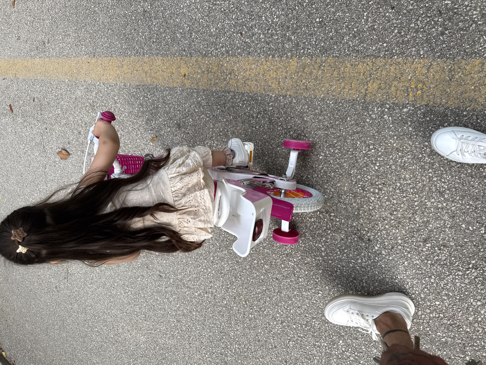 An overhead shot captures a child and an adult's feet on a grey asphalt or concrete path, marked by a faded yellow line stretching across the middle of the frame. The scene appears to be an outdoor area, possibly a sidewalk or park pathway, under natural daylight.
A young child, presumably a girl given her long, dark hair flowing over her back, is positioned horizontally across the lower left and center of the image. She is wearing a light-colored, possibly cream or beige, dress or top with patterned eyelet details. A small, star-shaped hair clip is visible in her hair near her head.
Next to the child, primarily in the mid-right of the frame, is a pink and white child's ride-on toy, resembling a tricycle or scooter with two prominent pink wheels and a white seat. The child's right hand is gripping the pink handlebar, and her right foot, clad in a white sneaker with frilly socks, rests on a pedal.
In the bottom right corner, and partially in the upper right, an adult's feet are visible, wearing clean white sneakers. One ankle shows a thin black anklet or band.
The image shows no discernible text or specific location markers beyond the general outdoor path, though the context provided indicates the location is Sandanski, Bulgaria. The activity seems to be a casual outdoor moment with the child on or near her ride-on toy.