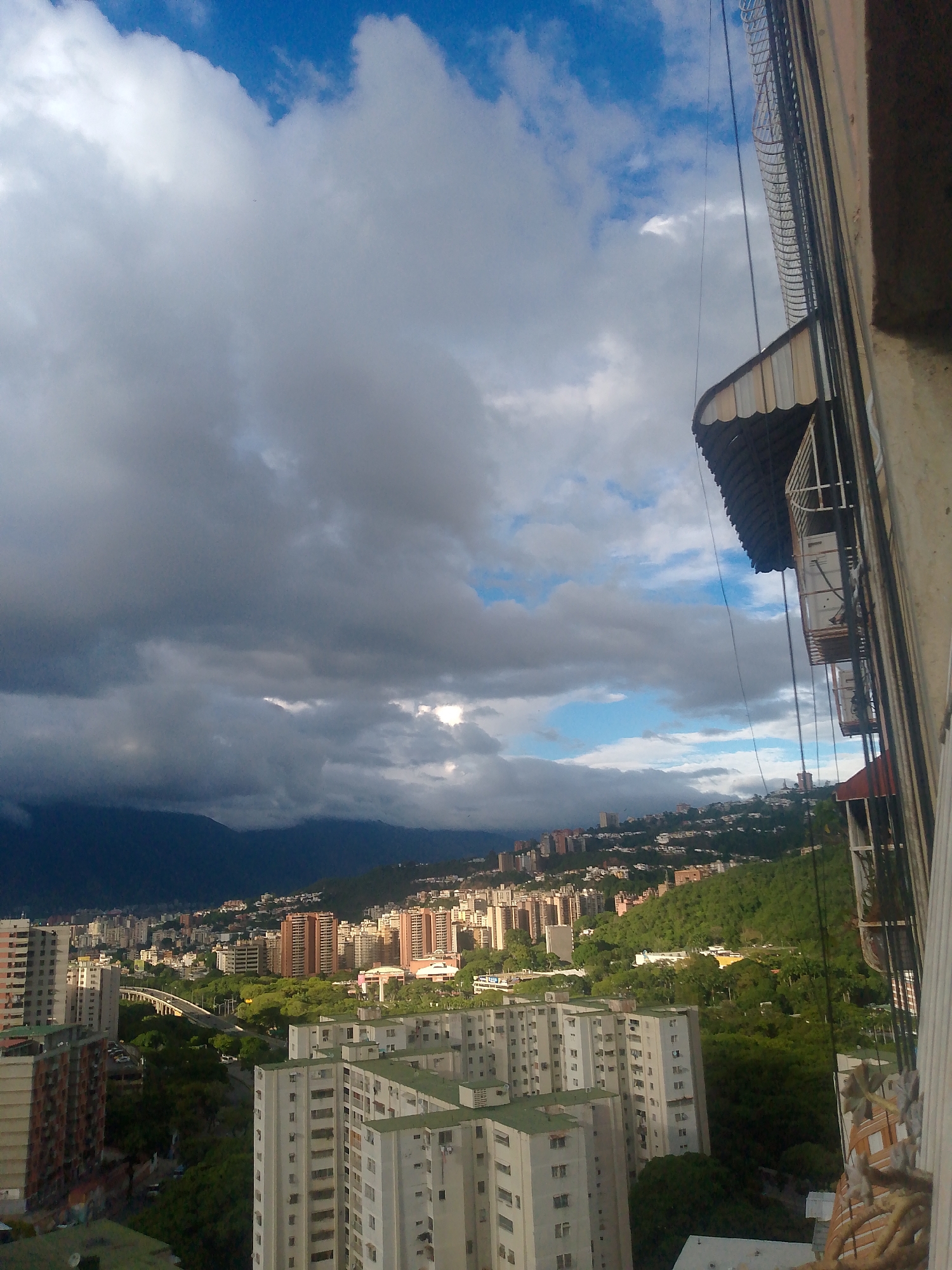 This is a high-angle, wide shot of a cityscape in Caracas, Venezuela, on a cloudy day. In the foreground, to the right, is a concrete balcony with a metal railing and awnings. The balcony extends out over the city.
The midground shows a dense urban environment with numerous residential buildings, mostly high-rises of varying architectural styles and colors, scattered across hills. Many of these buildings have a light-colored facade and green roofs. A multi-lane highway with an overpass is visible, winding through the trees and buildings. Lush green vegetation covers the hillsides, interspersed with more buildings, suggesting the city is built into the natural landscape. In the far background, dark, hazy mountains loom under a dramatic sky.
The sky is a mix of bright blue patches and large, dark, heavy clouds, indicating a potential for rain. The lighting suggests it is late afternoon, with sunlight occasionally breaking through the clouds, illuminating parts of the city and the mountainside. There are no people or discernible actions taking place within the frame. The overall impression is of a sprawling, densely populated city nestled within a mountainous region.