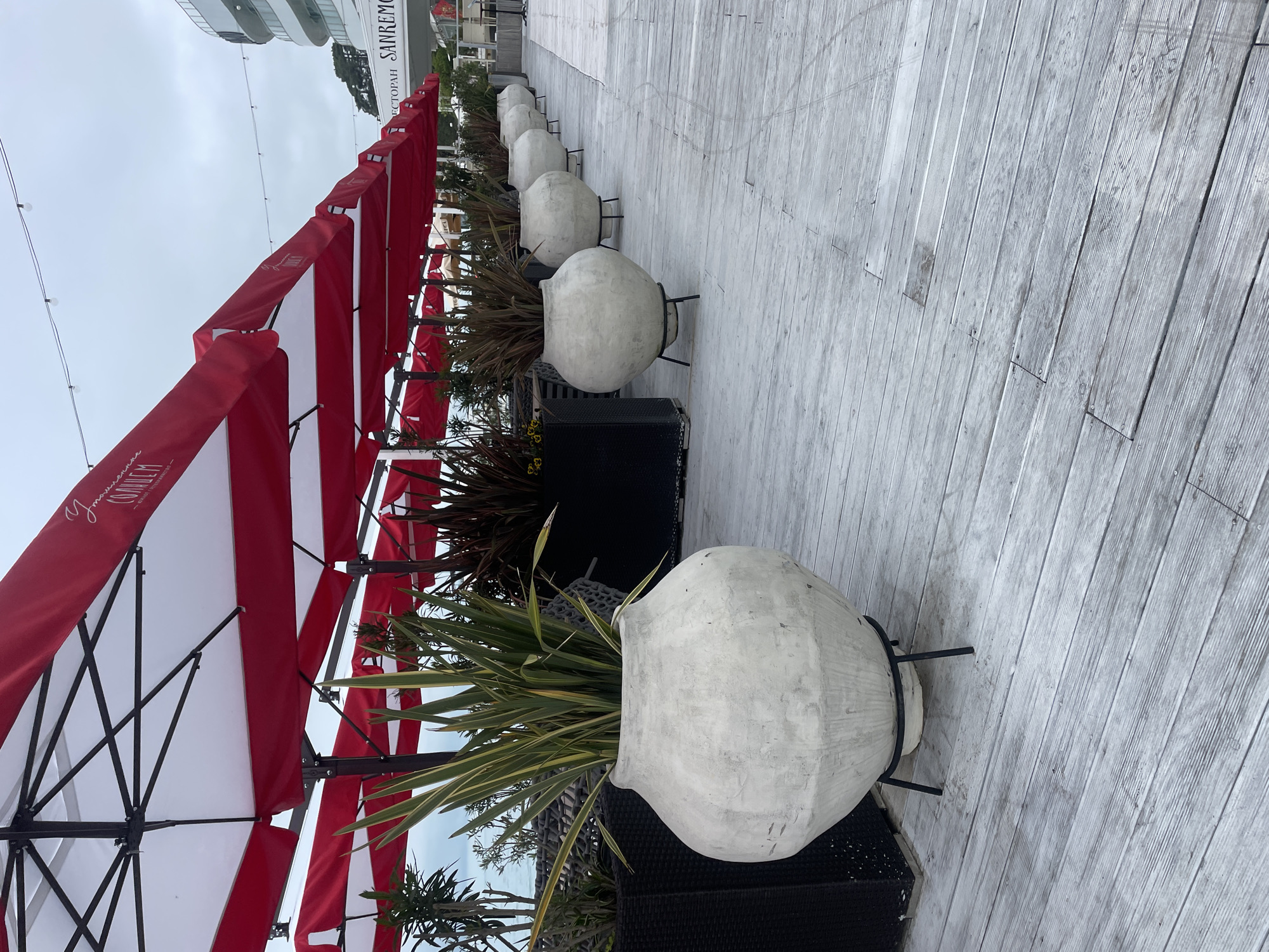 The image shows an outdoor dining area, likely a patio or boardwalk, with several round, white planters containing greenery. The planters are placed along a wooden deck, adjacent to a series of red and white striped umbrellas, typical of restaurant or cafe settings. The background features a modern building structure, indicating an urban setting. According to the writing on the edge of the building, the location is the city of Sochi, Russia. The environment appears to be well-maintained and indicative of a restaurant, cafe, or dining area along the sea.