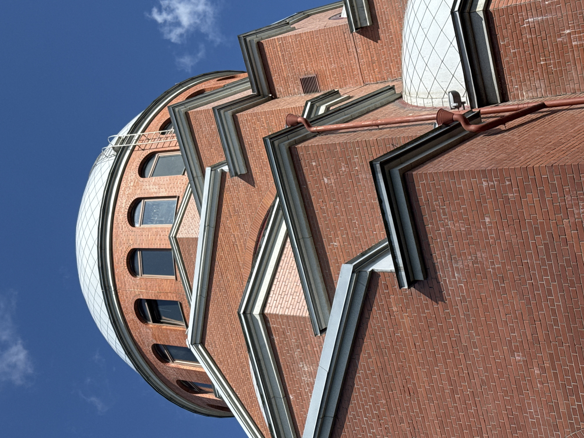 The image shows a low-angle, upward view of a brick building with a dome. The building is constructed of red bricks with white trim around architectural features like the windows and roof edges. The dome is silver and segmented. There are several arched windows on the curved brick section below the dome. A red downspout runs vertically along the side of the building, disappearing behind a corner. The sky above is a bright blue with a few wispy white clouds. The scene suggests an outdoor architectural shot. The provided context indicates the location is the city of Sinyavino, Russia, specifically mentioning the Great Choral Synagogue.
