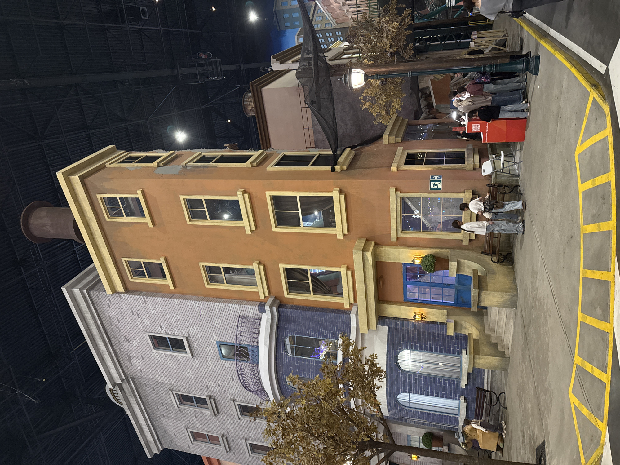 A vertical shot reveals a detailed scene of an urban environment, seemingly a movie set or themed area. The architecture is reminiscent of classic city buildings, with a mix of brick and stucco facades. The structures feature multiple windows, some with decorative moldings. There's a street-level entrance with a blue door and steps leading up to it. A mock street runs alongside the buildings, complete with yellow painted crosswalk lines, benches, streetlights, and trees with sparse foliage. A group of people stands together, interacting with what appears to be a booth or station.
Based on the architectural style, this setting does not appear to be located in Moscow, Russia. The architectural design is more in line with an American or European city than with the typical architecture of Moscow.