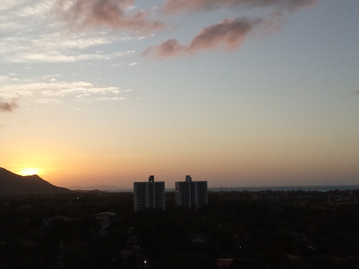 A wide-angle, elevated view depicts a sunset over the city of Juan Griego, Venezuela. The sky transitions from light blue with sparse white and pink clouds in the upper portion to a bright orange and yellow gradient near the horizon. The setting sun, a prominent orange disc, is positioned low on the far left, partially obscured by a large, dark mountain silhouette. In the mid-ground, two identically structured, multi-story buildings, light in color, stand erect against the illuminated sky. Below these structures and extending across the lower portion of the image, a dark, silhouetted urban landscape of trees and buildings is visible. The far right horizon line reveals a dark, calm body of water, indicative of the ocean. No individuals or specific activities are discernible within the frame.