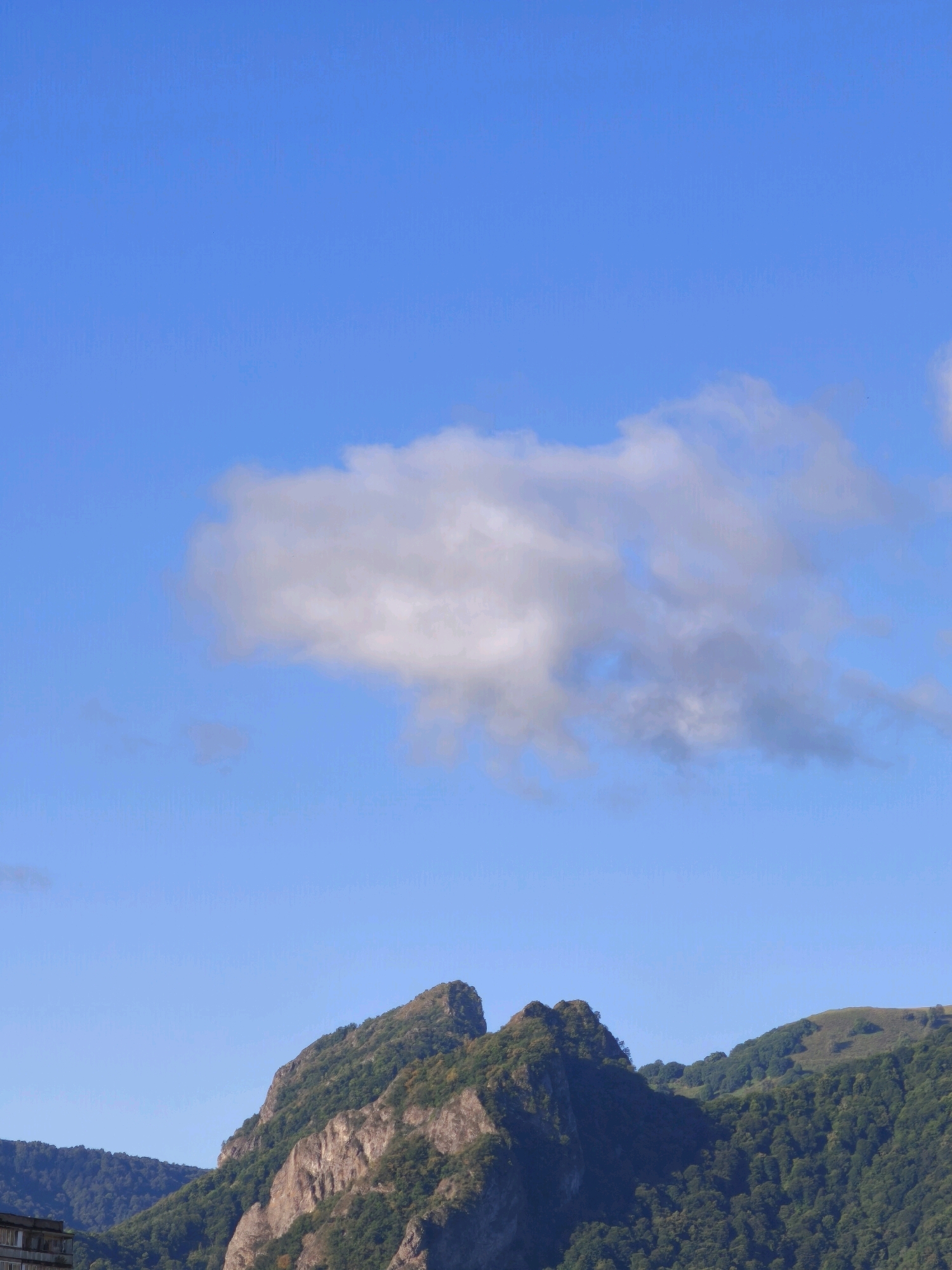 This image depicts a mountainous landscape under a clear blue sky with a few scattered clouds. In the foreground, on the left side, a building with a light-colored facade and visible windows is partially visible. The central focus of the image is a dramatic mountain peak with rugged, rocky slopes and patches of green vegetation. To the right of this peak, a gentler, rolling hill covered in dense green trees rises. The overall environment suggests a natural setting, likely on the outskirts or within a mountainous region such as Karachayevsk, Russia, which is known for its scenic mountain vistas. No people or specific actions are captured in the frame.