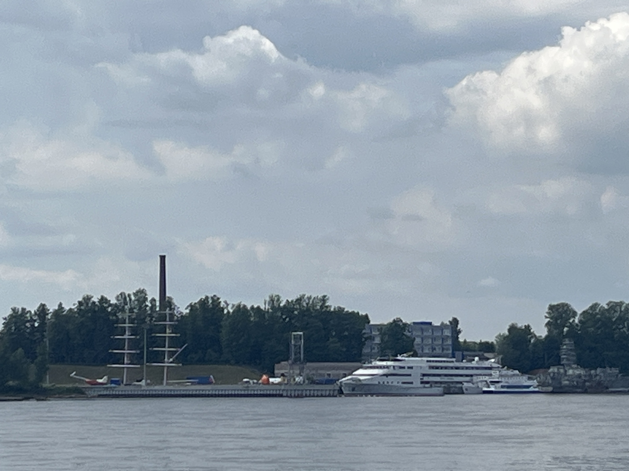 The image shows a wide, calm body of water in the foreground, likely a river or bay. In the middle ground, a dock area is visible, lined with trees. On the dock, there are several objects of interest. To the left, a tall, narrow smokestack stands out against the treeline. Next to it, a replica of a sailing ship with multiple masts is displayed. Further to the right, a modern, multi-decked white passenger ship is docked. Adjacent to the passenger ship, there are smaller vessels. The background consists of a densely wooded area, with buildings of varying heights peeking through the trees. The sky is overcast with scattered white clouds. The image appears to be taken from a boat, looking towards the shore. The scene depicts a harbor or waterfront area in a populated locale, possibly a tourist destination or a working port, in the city of Imeni Sverdlova, Russia.