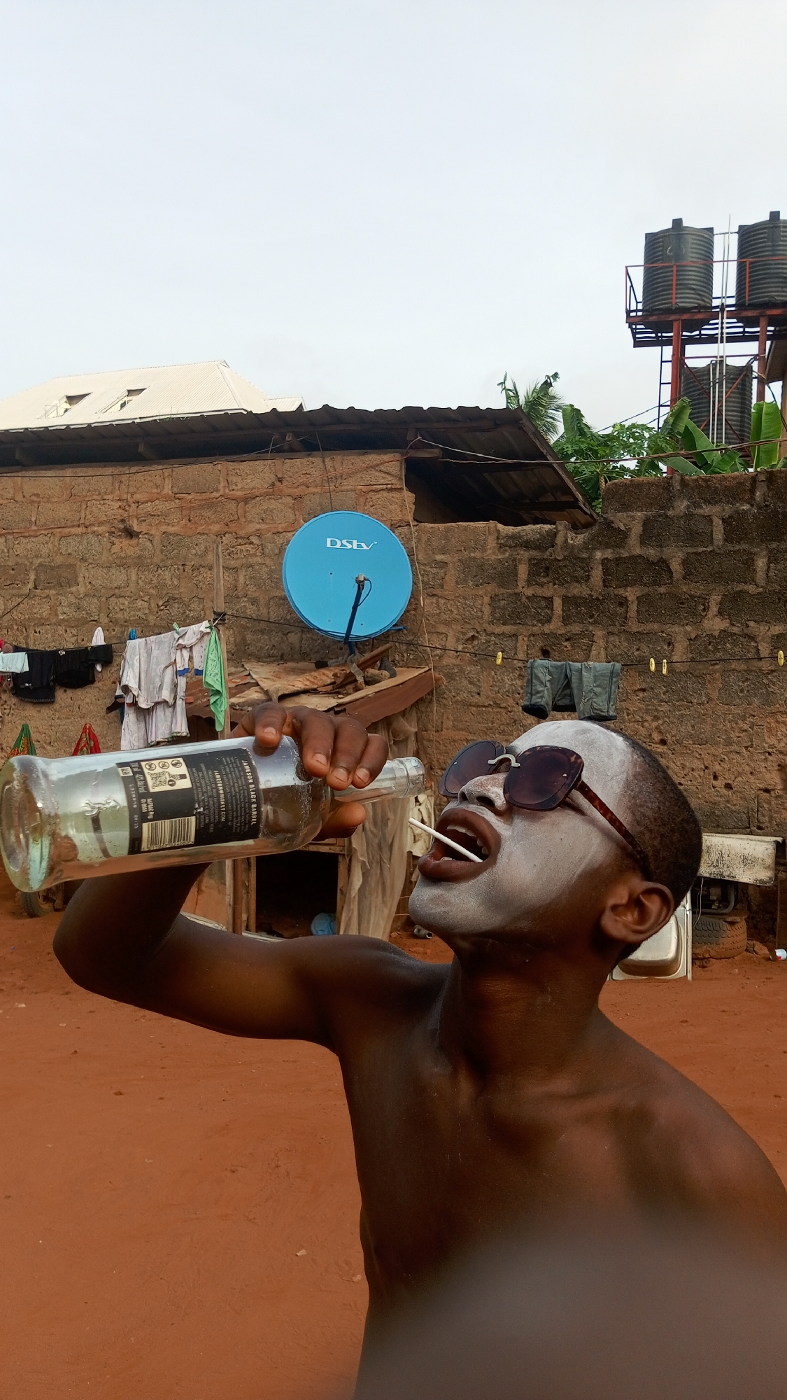 A shirtless individual with white face paint and sunglasses is drinking from a clear glass bottle. A small white stick protrudes from their mouth. Behind the individual, a weathered brick building is visible, with clothes hanging on a line. A blue DStv satellite dish is mounted on the wall. In the background, water tanks are perched on a metal scaffolding. The ground is a reddish-brown dirt. The location is in an undefined area, city of Unknown, Unknown.