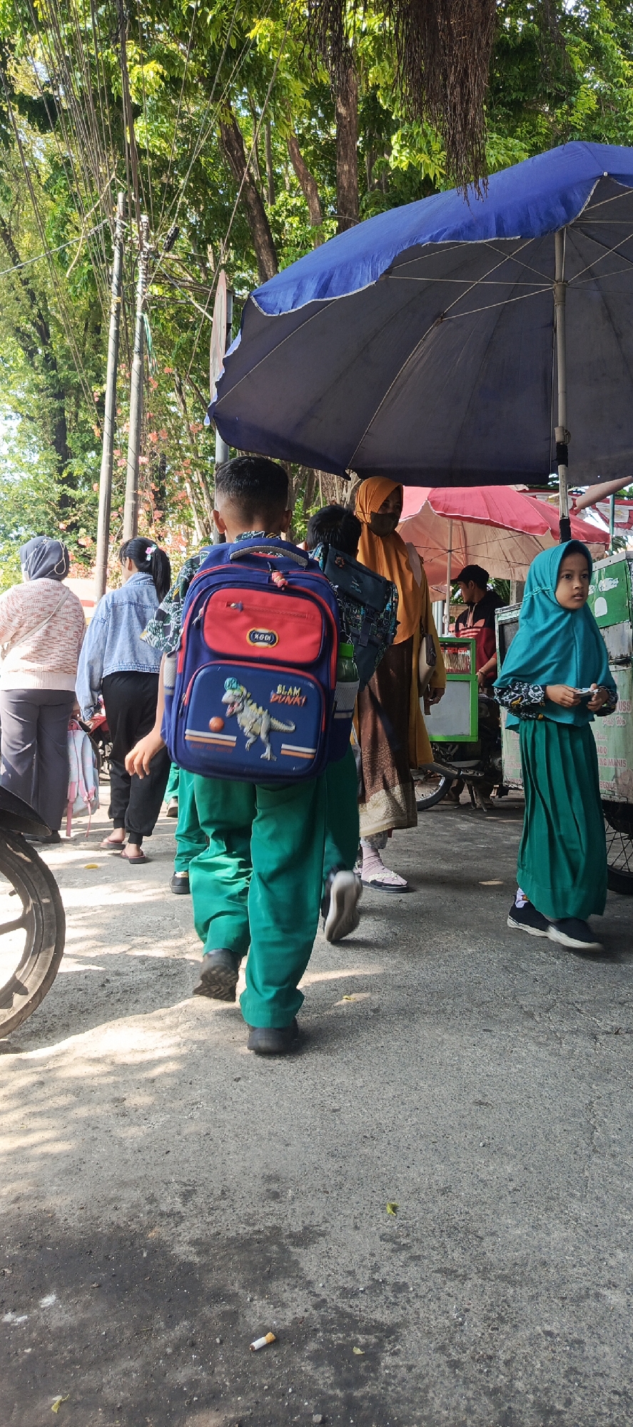 The image depicts a street scene in Krian, Indonesia, with several individuals present. In the foreground, a young boy wearing a blue and red backpack with a dinosaur design and green pants is walking away from the camera. To his left, a woman in a grey hijab and patterned sweater is also walking away. To the boy's right and slightly behind him, another child, possibly a girl, is wearing a turquoise hijab and green dress, looking towards the right. Further in the background, other people are visible, including a woman in an orange hijab. The environment includes trees overhead, a large blue umbrella, and a red umbrella. The ground is paved with asphalt, and a used cigarette butt is visible on the pavement in the lower right portion of the image. A portion of a motorcycle wheel is visible on the far left.