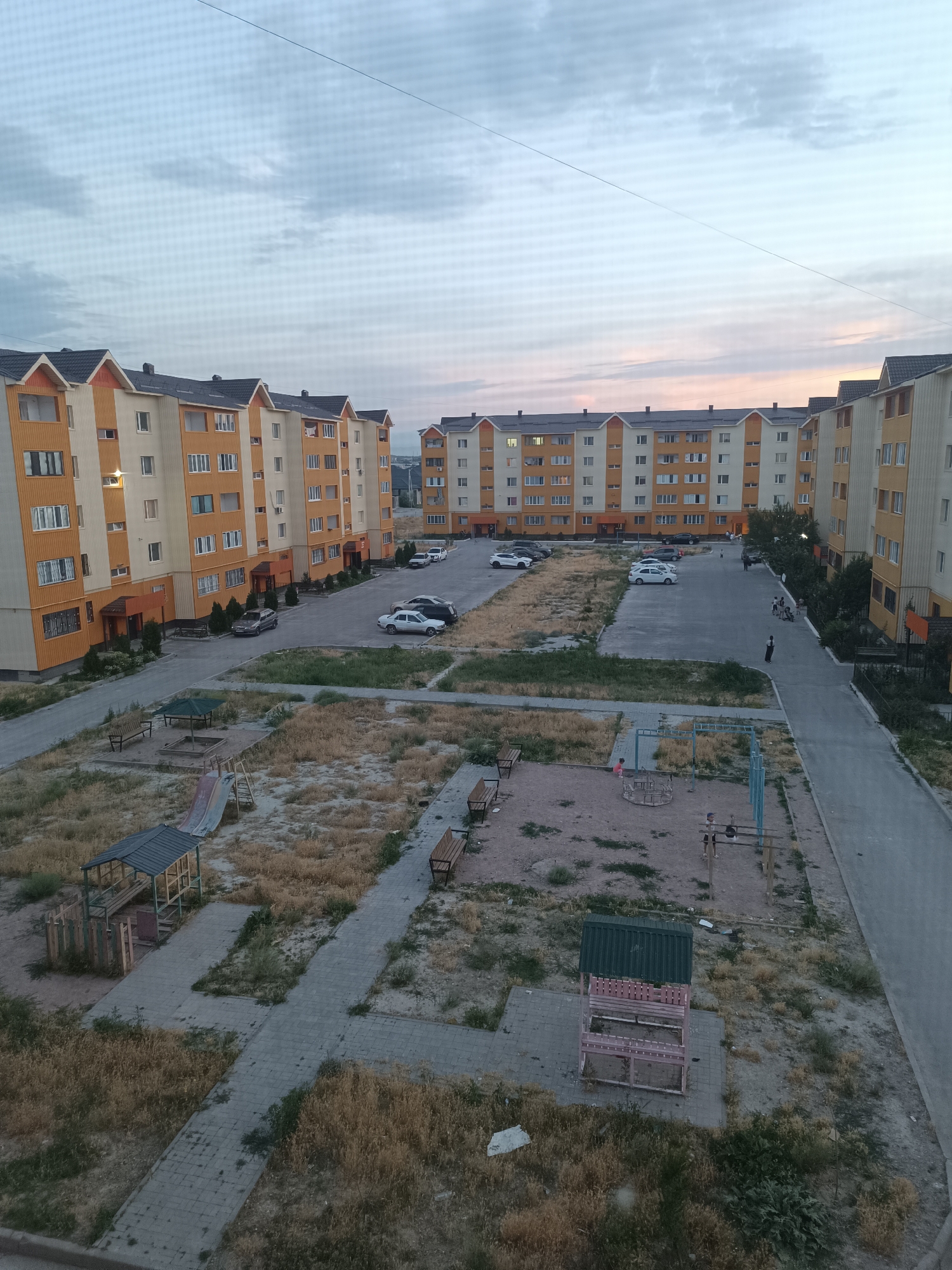 This is a high-angle, medium shot of an apartment complex courtyard in Taraz city administration, Kazakhstan, taken at dusk. The courtyard is paved with grey interlocking bricks and has patches of dry, yellowed grass. Several apartment buildings, painted in orange and cream, form the perimeter of the courtyard. In the foreground, a playground area features a red and silver slide, a sandbox with a green roof structure, and a metal swing set. Benches are scattered throughout the courtyard. Several cars are parked in designated spots. In the background, more apartment buildings are visible, along with a power line stretching across the sky. The sky is a pale blue with streaks of pink and orange from the setting sun, and there are scattered clouds. A few people are visible in the distance walking or standing in the courtyard. The scene appears to be a typical residential area.