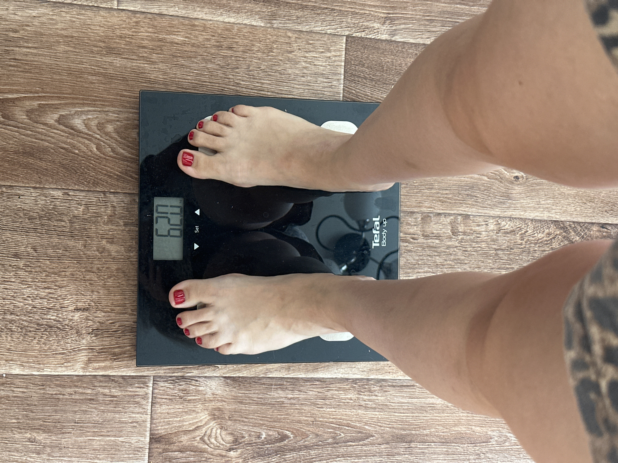 The image shows a person's feet and lower legs standing on a black digital bathroom scale. The scale displays the number "62.0". The scale is placed on a wooden floor with visible grain. The person's toenails are painted red. The image was taken in a residential interior. There is no information in the image to determine the location in the city of Novoaltaysk, Russia.