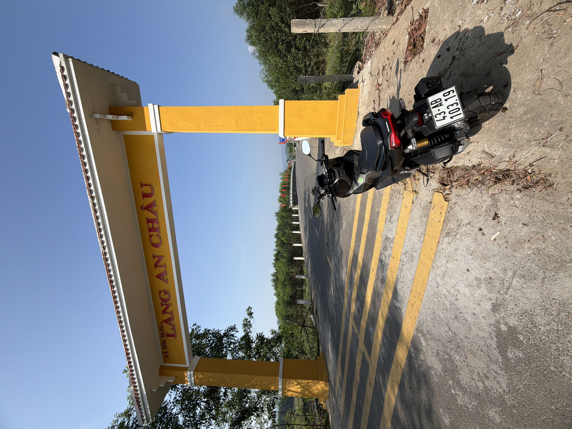 The image shows a motorcycle parked near a monument that reads "Làng An Châu" in the city of Đà Nẵng, Vietnam. The monument is yellow with white trim. The motorcycle, a black Honda ADV150, is parked partially on the paved road shoulder. The license plate number is 43-A8 103.19. The road is marked with yellow stripes. Trees and bushes are visible in the background. The weather appears sunny.