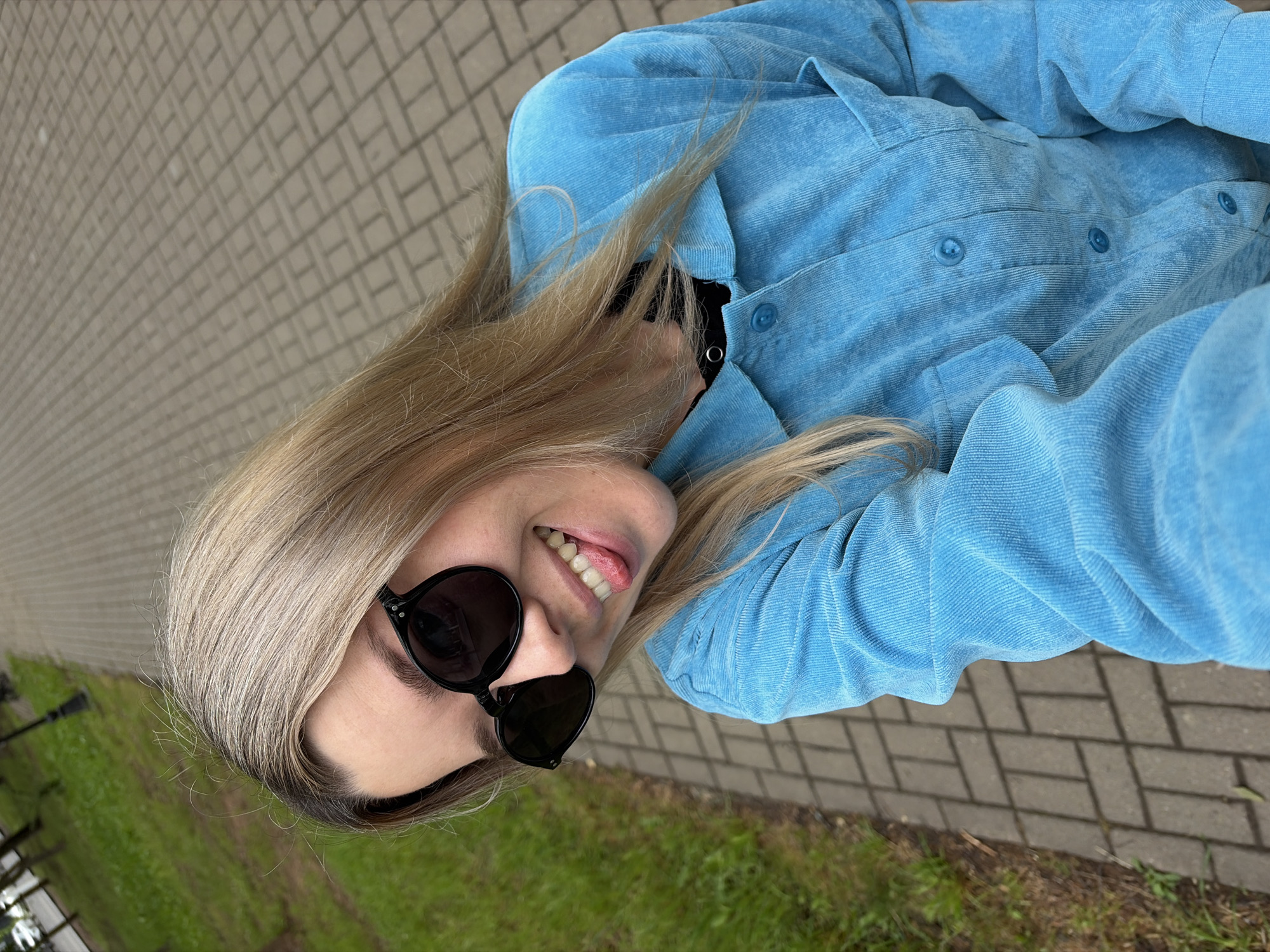 The image features a young woman with long, blonde hair and darker roots, captured in a playful moment outdoors in Chaikovsky, Russia. She is wearing dark, round sunglasses and a bright blue jacket made of a textured, corduroy-like fabric, with visible buttons down the front. Her expression is cheerful, characterized by a wide smile and her tongue slightly extended.
The photograph is taken from a high angle, looking down at the woman. Her upper body is largely framed by her hair and the distinct blue jacket. The setting is split between a light-colored paved path with a herringbone brick pattern on one side, and a strip of vibrant green grass on the other. Faint dark shapes, possibly lampposts or trees, are visible in the distance across the grass.
The lighting suggests it is daytime with fair weather. The woman's relaxed pose and happy expression convey a lighthearted mood. There is no visible text in the image.