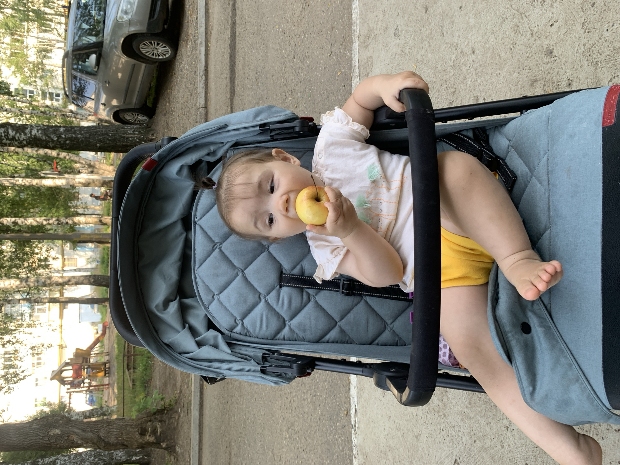 A young child sits in a stroller, holding a yellow apple to their mouth. The child wears a light-colored shirt with ruffles on the sleeves and bright yellow shorts. The stroller is teal-colored with a padded seat and a black safety bar. In the background, trees line a paved area. A silver SUV is parked on the right side of the frame, and a playground structure with a slide is visible behind a fence on the left. The scene appears to be in a residential area, likely in the city of Chaikovsky, Russia, given the architectural style of the building and the vegetation.