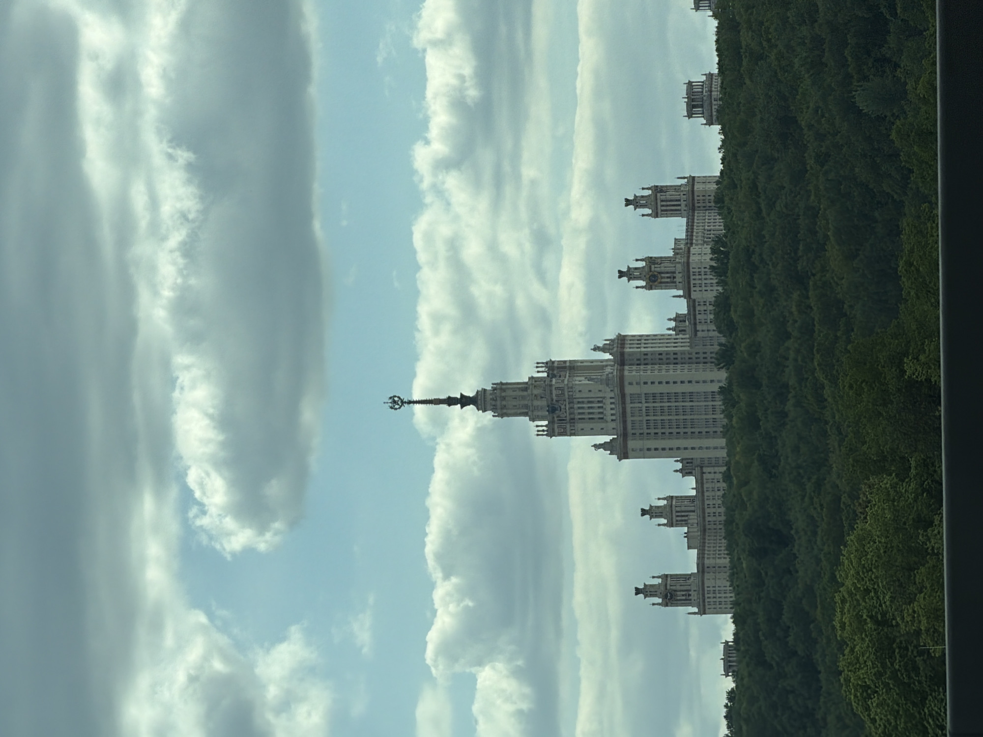 The image shows a tall, ornate building, likely a university structure, amidst trees and clouds. The building is the Moscow State University, one of the Seven Sisters skyscrapers. The architecture is Soviet-era Stalinist style with a central tower topped by a spire. The surroundings include dense green trees and a sky filled with cumulus clouds.