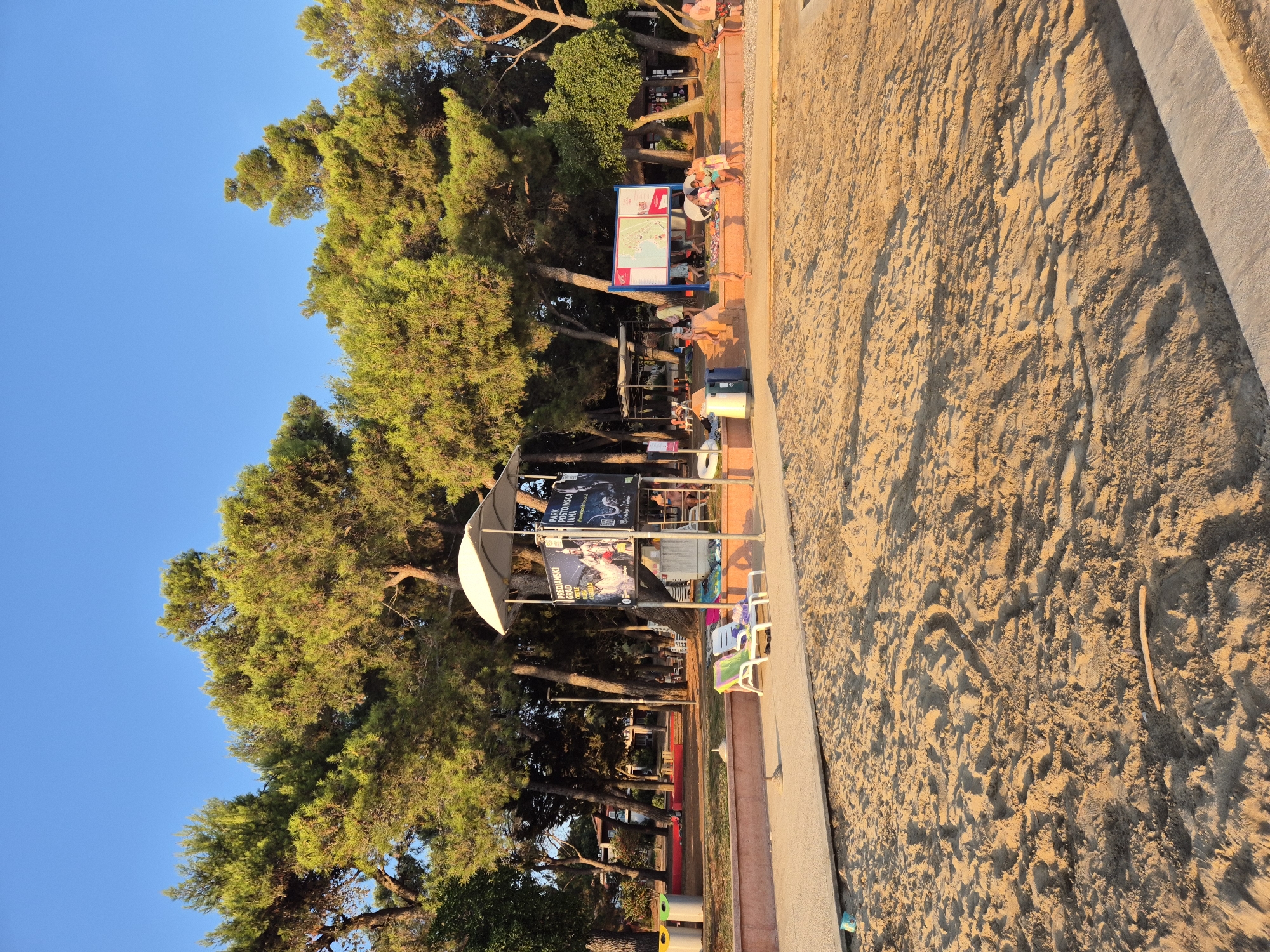 The image depicts a sandy beach area with lush green trees and a clear blue sky. In the background, there is a lifeguard tower with posters displayed on its sides. Several people are visible, some relaxing on lounge chairs, while others are walking or sitting on the sand. There are also amenities such as trash cans and a map display. The scene appears to be in Katoro, Croatia, likely a public beach or recreational area. The lighting suggests it is either early morning or late afternoon.
