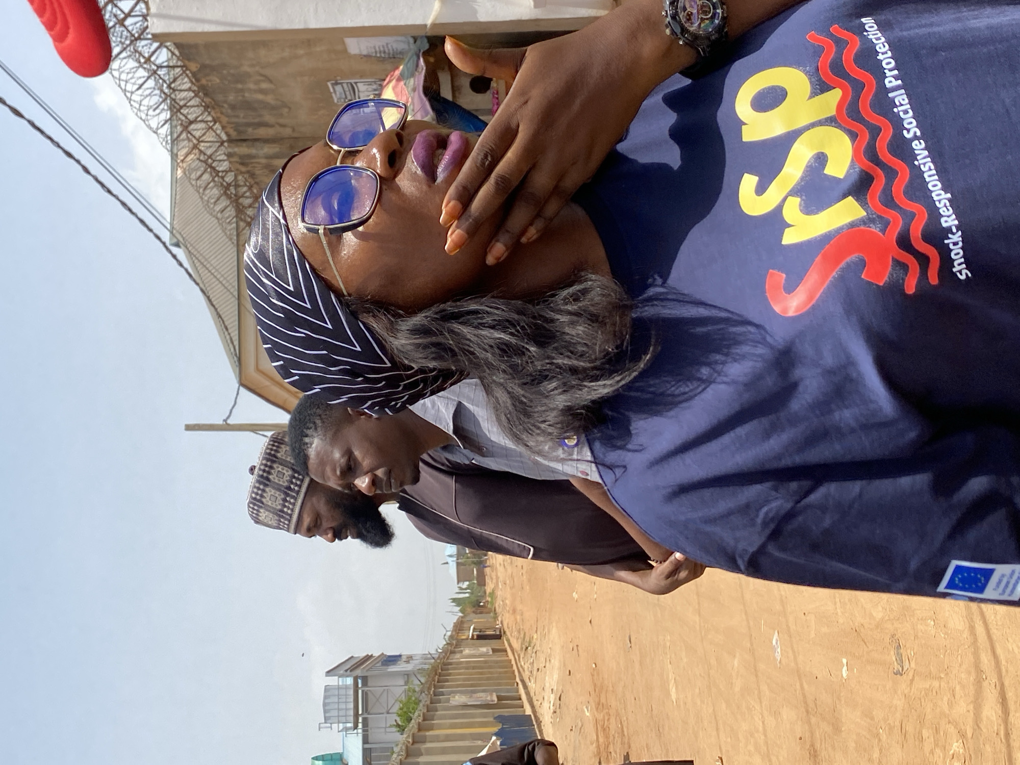 The image shows a medium shot of a group of people in what appears to be Sabon Tasha, Nigeria. In the foreground, a woman with dark skin, sunglasses, and a striped headscarf is looking upwards and wearing a dark blue shirt with the acronym "SYSP" and the phrase "Shock-Responsive Social Protection" printed on it. Her lips are painted a dark purple color, and she is wearing a watch on her left wrist. In the background, there are two men standing close together. One of the men has a beard and is wearing a patterned hat. The environment includes buildings, fences with barbed wire, and a dirt road, suggesting an outdoor location in Sabon Tasha. The European Union flag is on the bottom of the blue shirt.