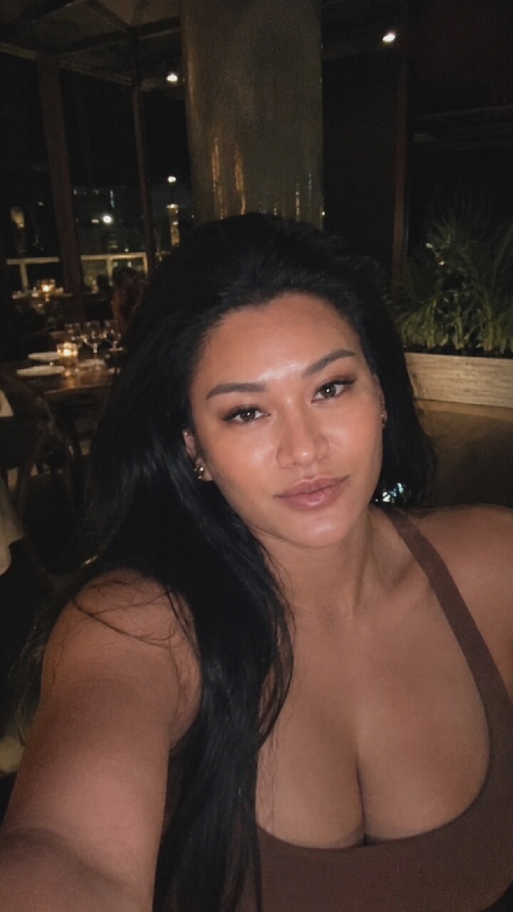 A woman with long, dark hair and light skin is pictured indoors, likely in a restaurant or lounge, as suggested by the blurred background featuring tables, chairs, and ambient lighting. She is wearing a brown top that reveals her décolletage. The woman is looking directly at the camera, with her head slightly tilted to her left. Her makeup includes defined eyebrows, eyeliner, and lipstick. The lighting in the image is dim, creating a sophisticated and intimate atmosphere. There is no indication of the location being in Moscow, Russia.