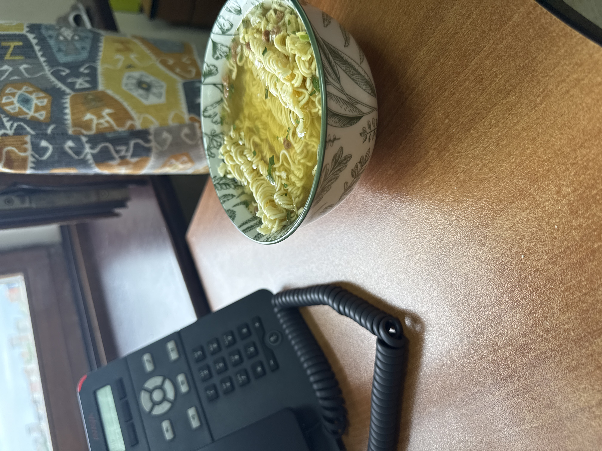 This media file shows a close-up, angled shot of a bowl of instant ramen noodles with broth, sitting on a wooden desk. The bowl has a green and white floral pattern. To the right of the bowl, a black corded phone with a keypad and a small LCD screen is visible. The background includes a blurry patterned fabric, likely upholstery, and a window with a view that is out of focus. The scene appears to be indoors, possibly in an office or home setting, with artificial lighting. No people are visible, and the time of day and weather are not discernible. There is no visible text in the image. The overall impression is a casual meal break at a desk.