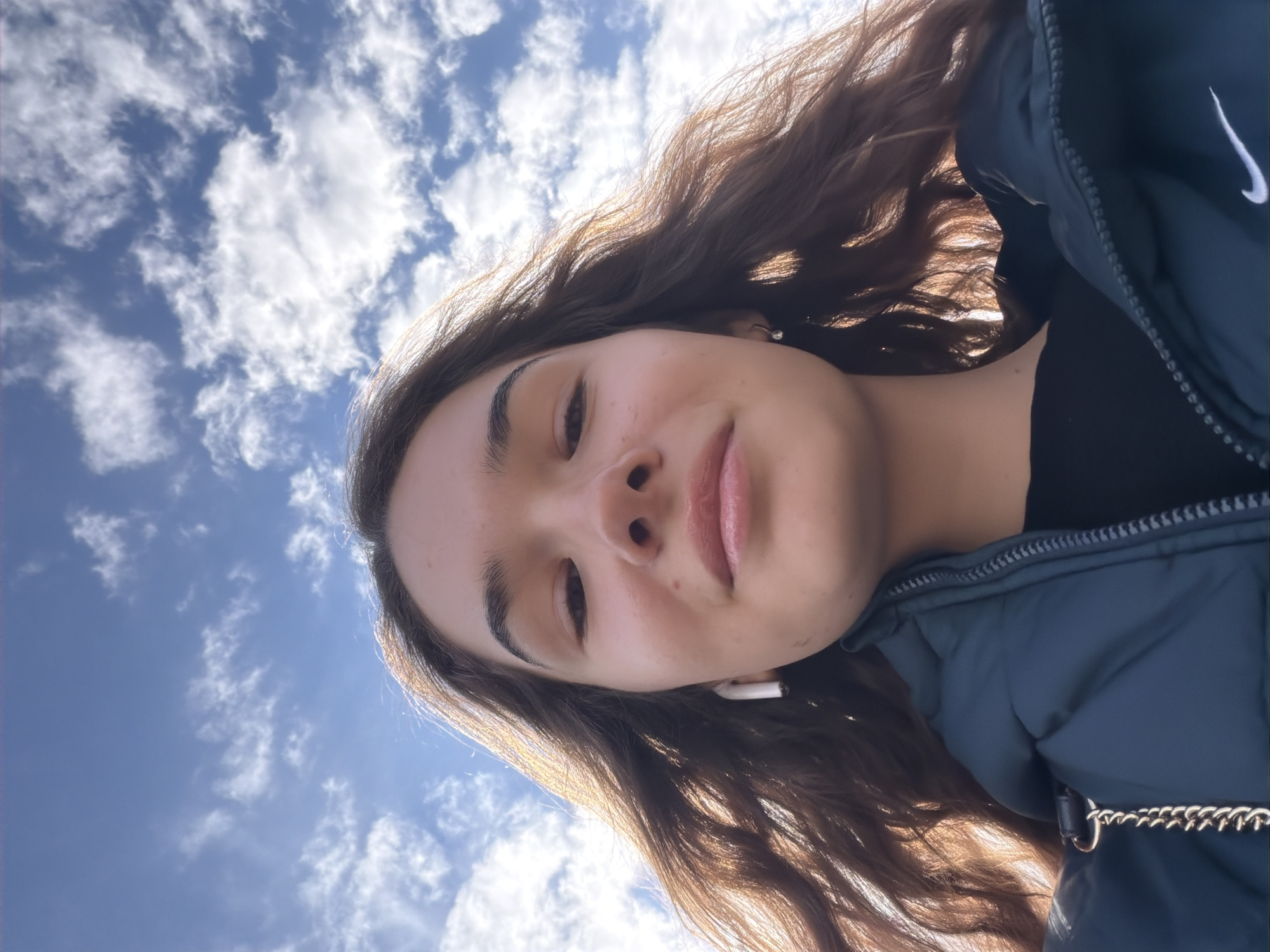 A low-angle medium shot captures a young woman in Sofia, Bulgaria. The sky is blue with scattered white clouds visible in the background. The woman has brown hair and is wearing a dark blue jacket with a white Nike swoosh on the shoulder. An earbud is visible in her ear. The context for the location of the subject is not provided.