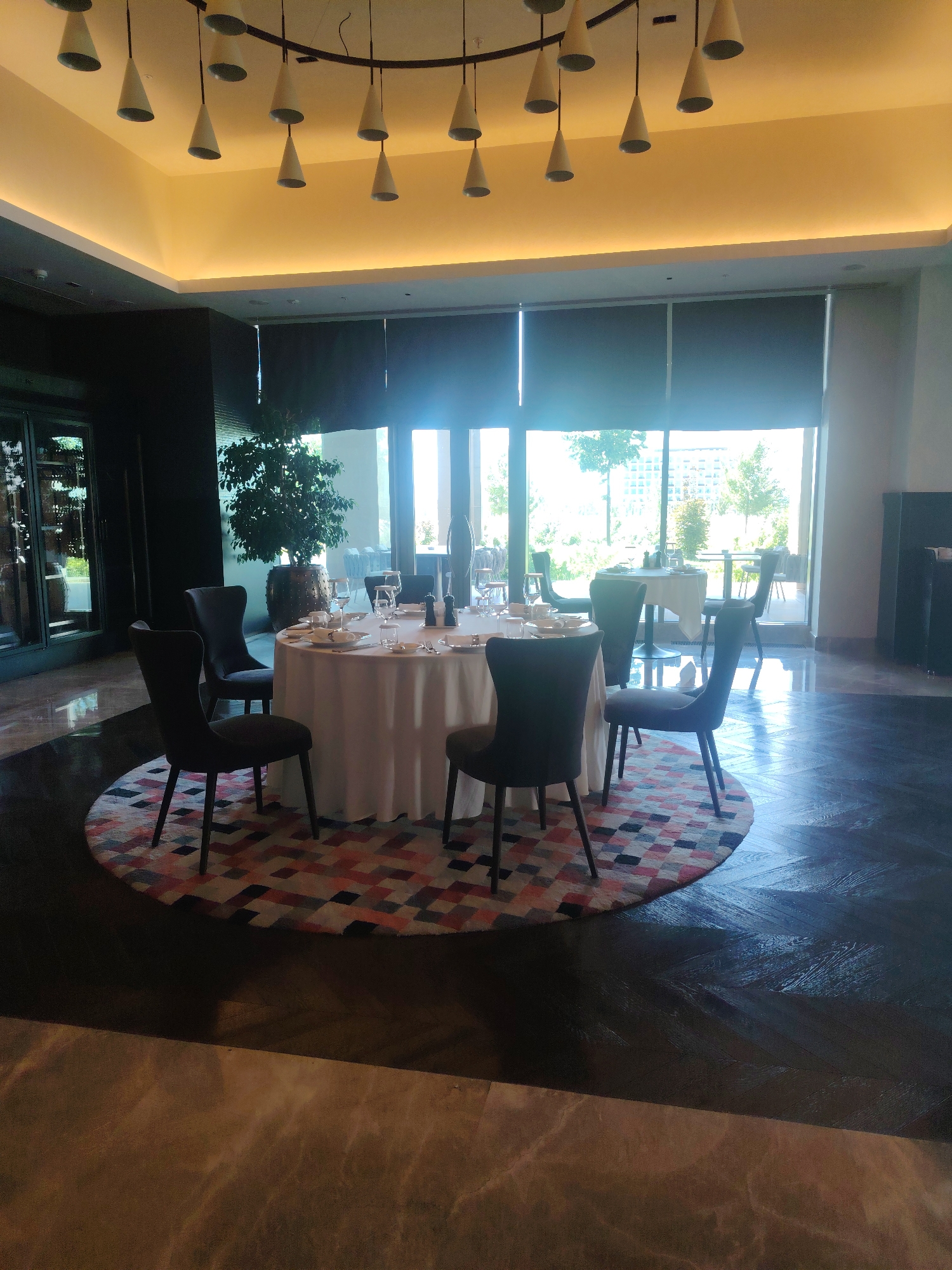 An interior scene shows a round dining table set for a meal in what appears to be a restaurant or event space. The table is covered with a white tablecloth and features multiple place settings, including plates, glasses, and cutlery. Two salt and pepper shakers are also visible on the table. Surrounding the table are four dark upholstered chairs. The table and chairs are positioned on a circular rug with a geometric pattern of red, pink, black, and white squares. The floor is dark wood with a parquet pattern. In the background, large windows offer a view of greenery and buildings outside, suggesting an urban setting. Several pendant lights hang from the ceiling, emitting a warm glow. A tall potted plant is situated to the left of the table. The overall impression is one of a formal and elegant dining area. This location is in the city of Konigil, Uzbekistan.